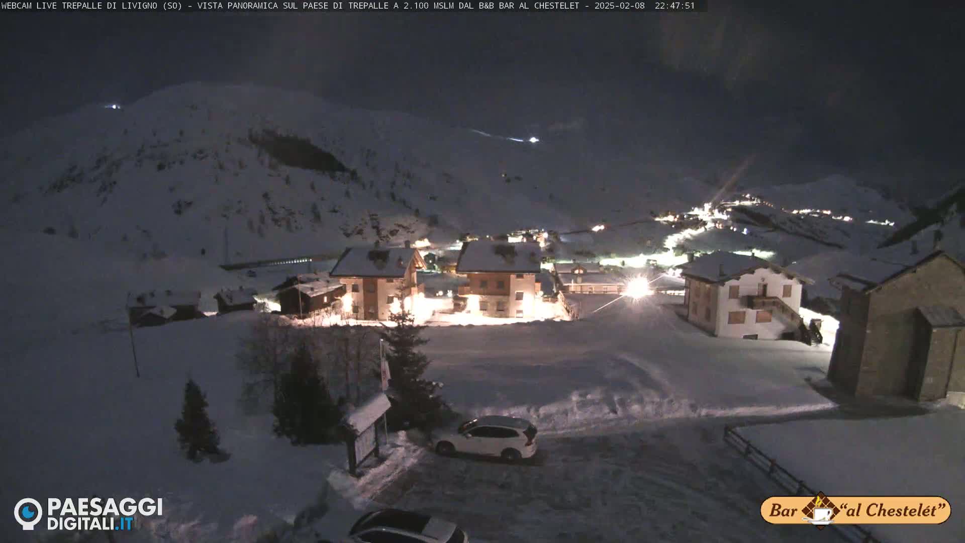 A snow-covered village is illuminated at night, nestled among snow-covered hills under a dark sky.