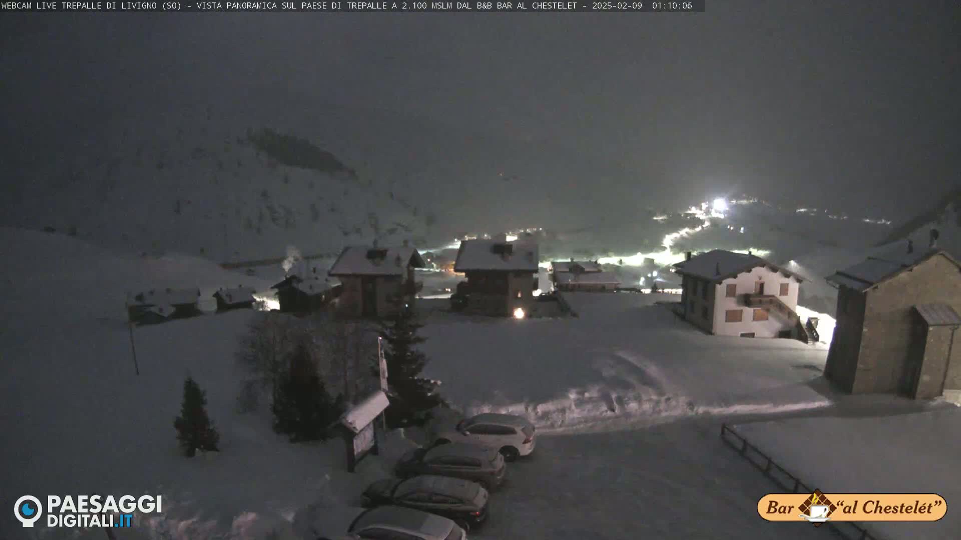 A snow-covered village at night is illuminated by lights from buildings and vehicles, with several cars parked in a snowy lot.