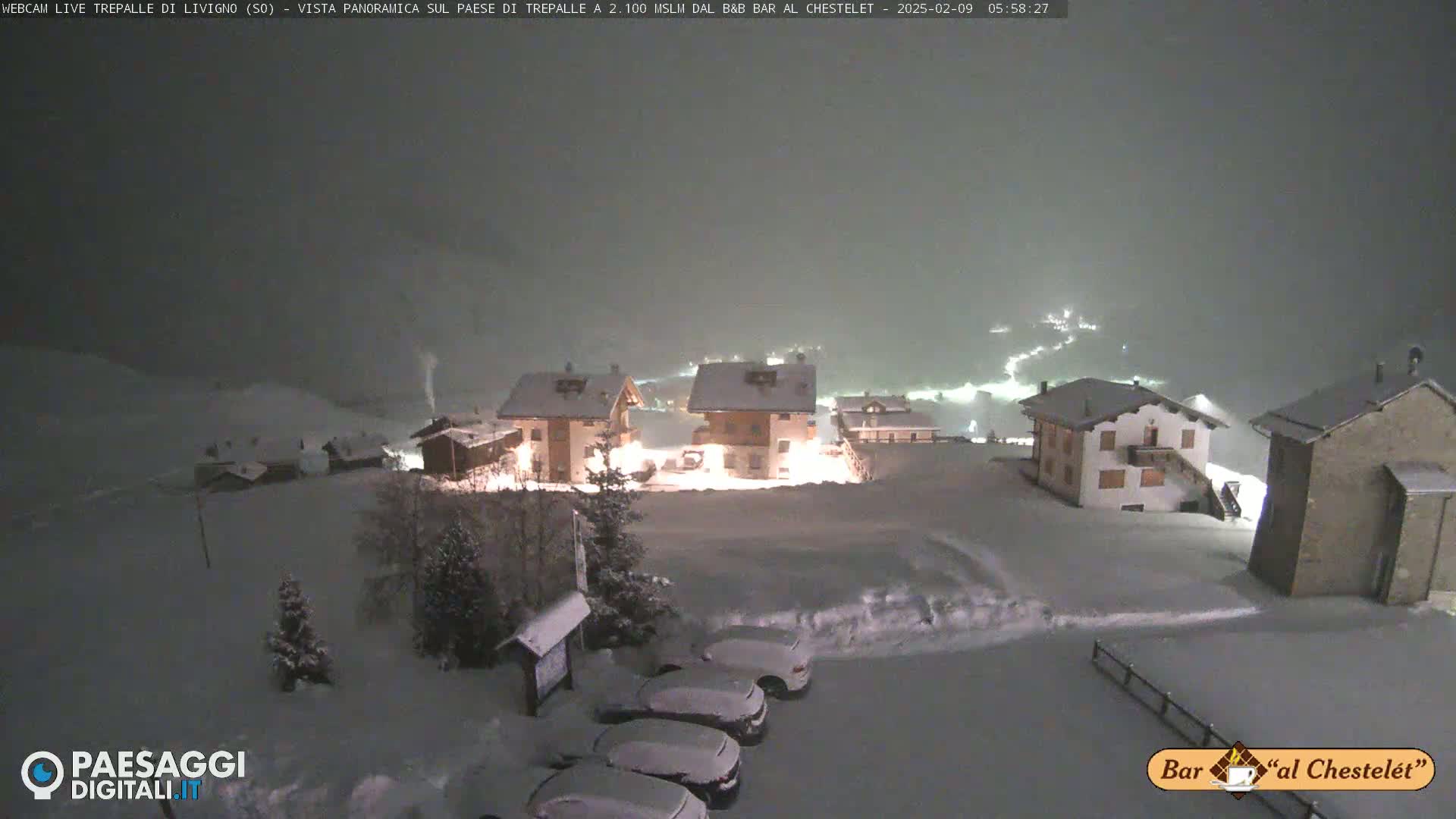 Snow-covered buildings are illuminated at night during a snowfall.