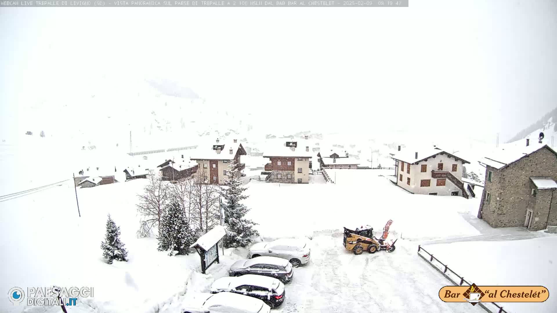 A snow-covered village scene shows several buildings, parked cars, and a small snowplow in the midst of a snowfall.