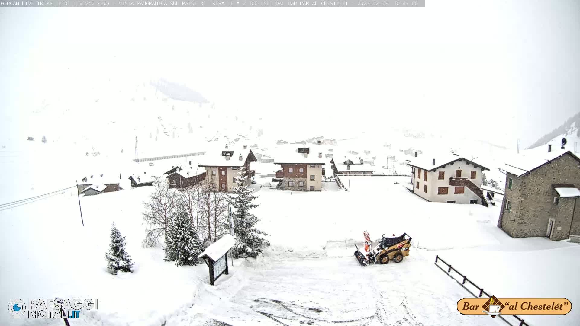 A snow-covered village is seen from an elevated vantage point, with a small snowplow clearing a path in the foreground during a snowstorm.