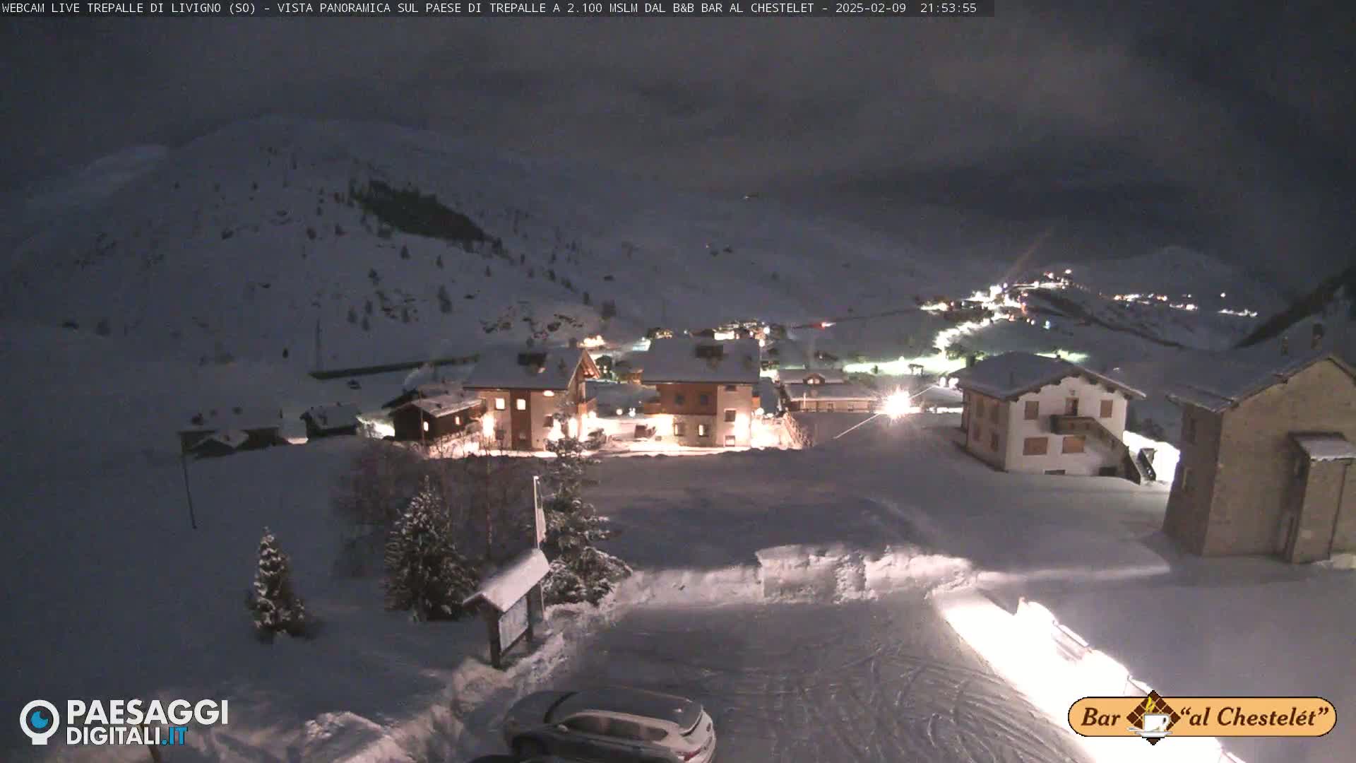 A snow-covered village at night is illuminated by lights from buildings, under a dark, overcast sky.