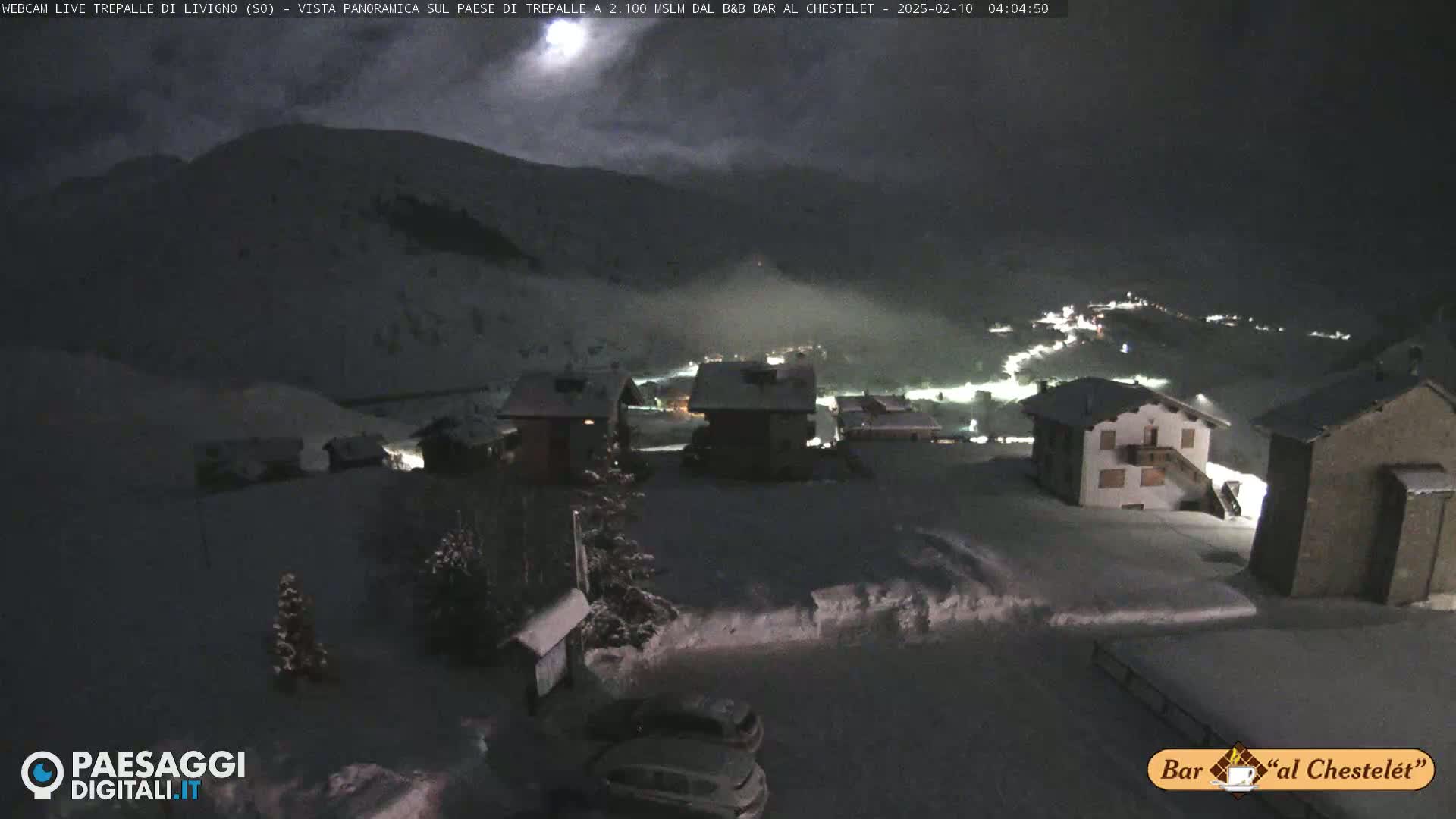 A snow-covered village at night is illuminated by scattered lights, with a mountainous backdrop under a dark, mostly cloudy sky.