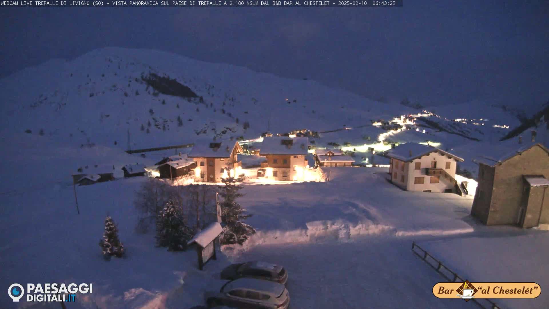 A snow-covered village at night, illuminated by lights from buildings, sits on a hillside under a dark, twilight sky.