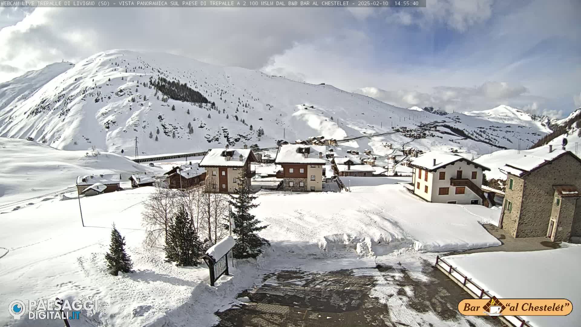 A snow-covered village nestled in a valley surrounded by snow-capped mountains under a partly sunny sky.