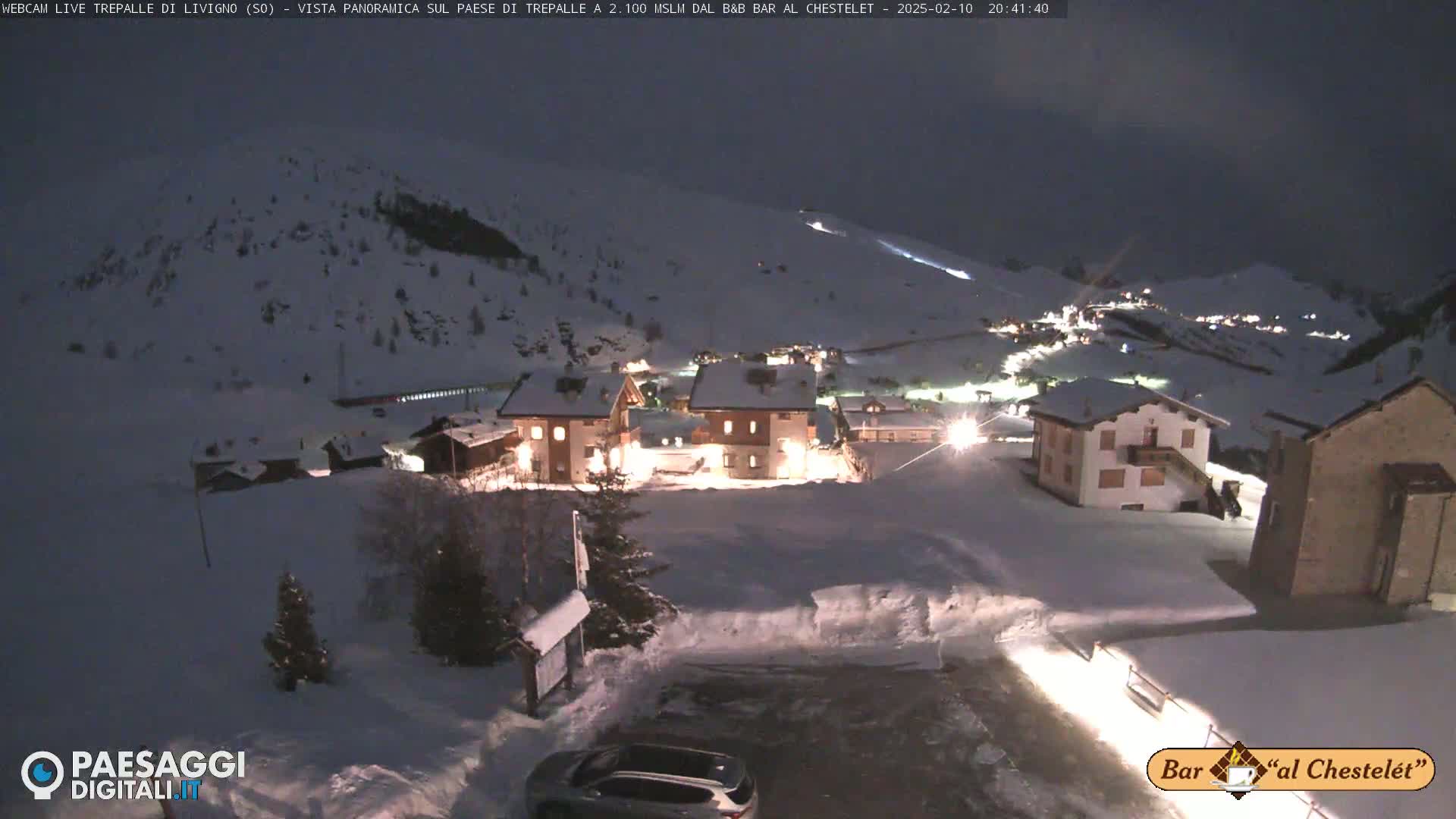A snow-covered village at night, illuminated by lights from buildings, is nestled on a hillside under a dark sky.