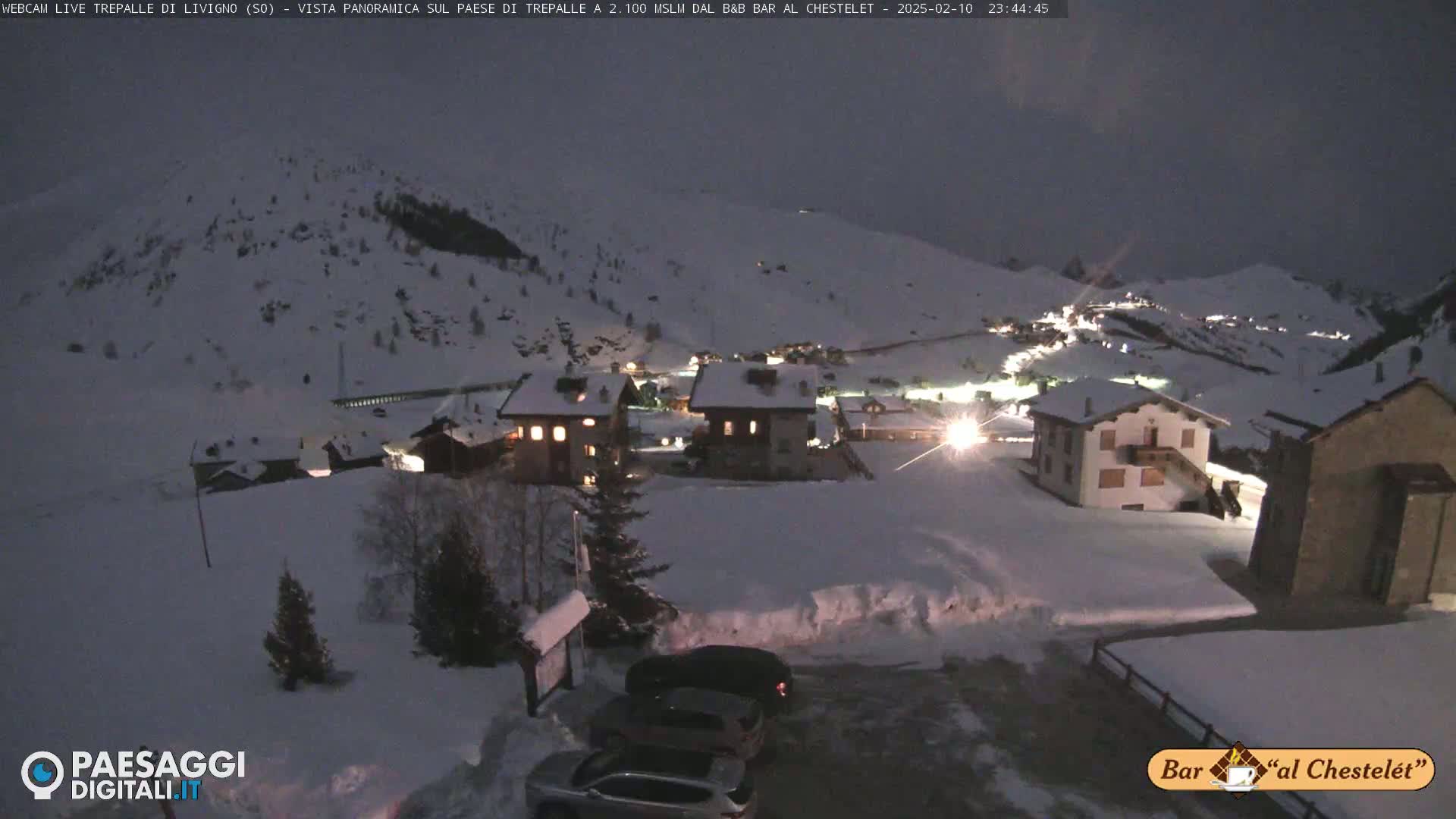 A snow-covered village at night, illuminated by scattered lights, is nestled in a mountainous landscape under a dark sky.