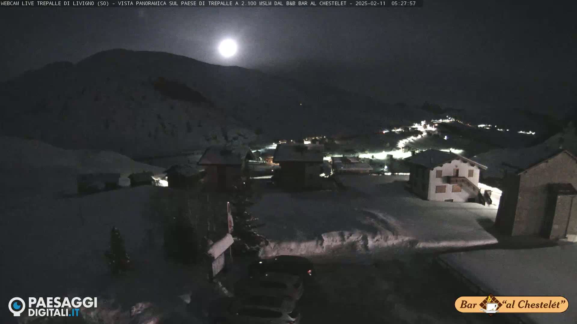 A bright moon illuminates a snow-covered village nestled in a mountainous region at night.