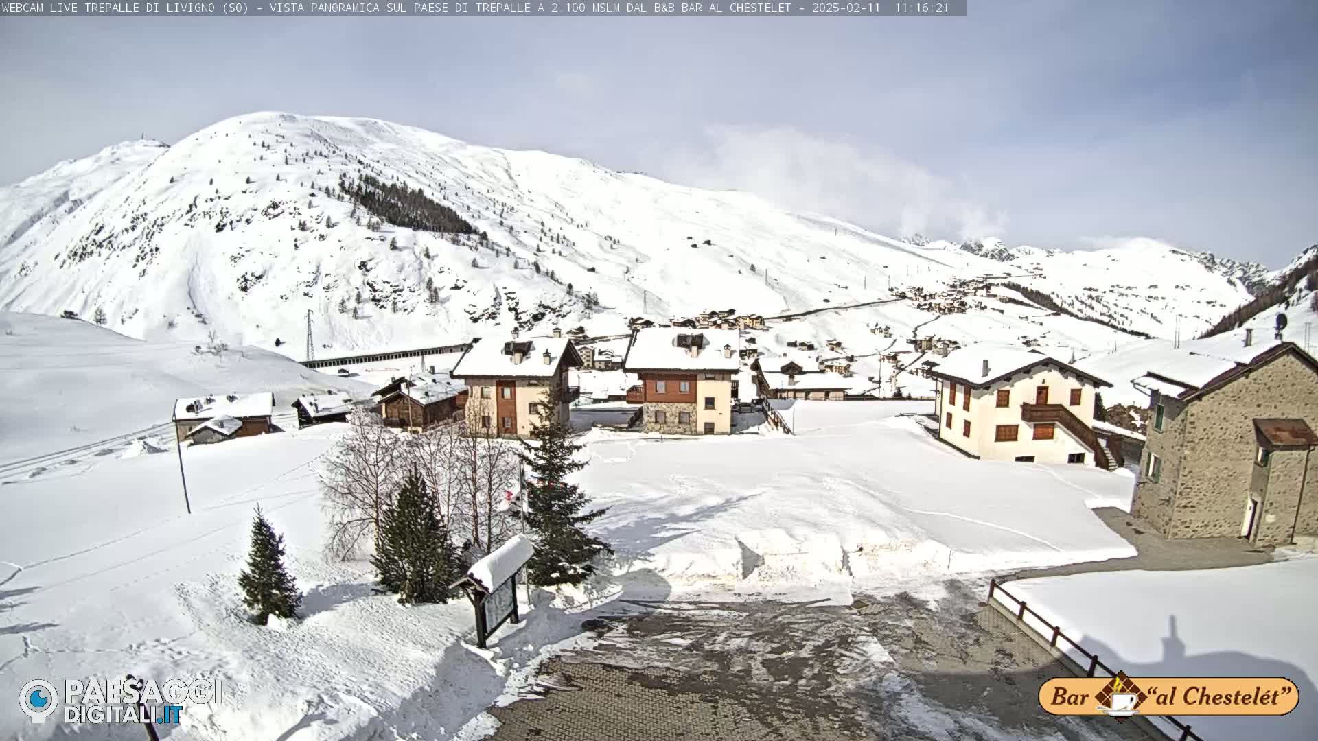 A snow-covered village nestled in a valley, with snow-capped mountains in the background under a mostly sunny sky.
