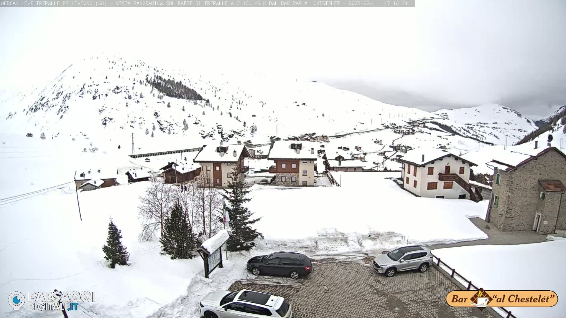 A snow-covered village nestled in a mountainous region under an overcast sky, with several cars parked in a lot.