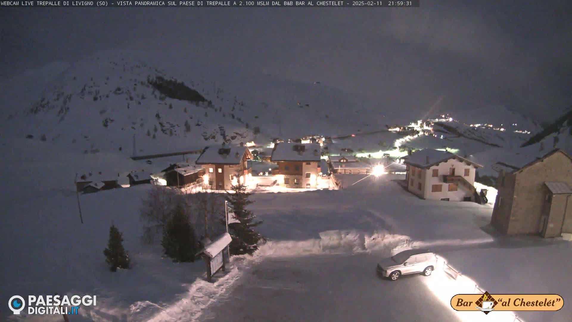 A snow-covered village at night, illuminated by lights from buildings, with a car parked in the foreground under a dark sky.