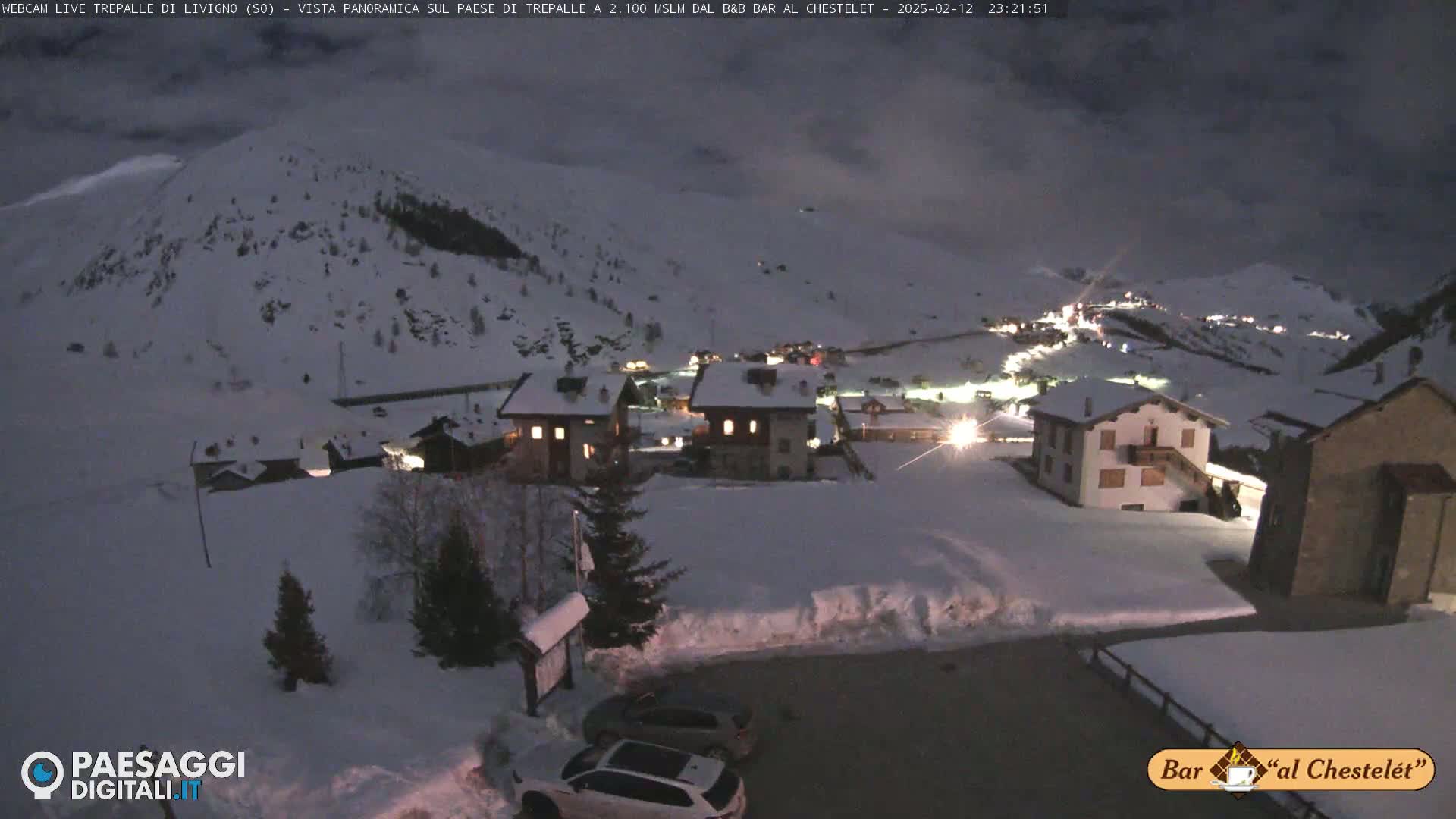 A snow-covered village at night is illuminated by dim lights, with cars parked in a snowy lot in the foreground and hills blanketed in snow in the background under a dark, cloudy sky.