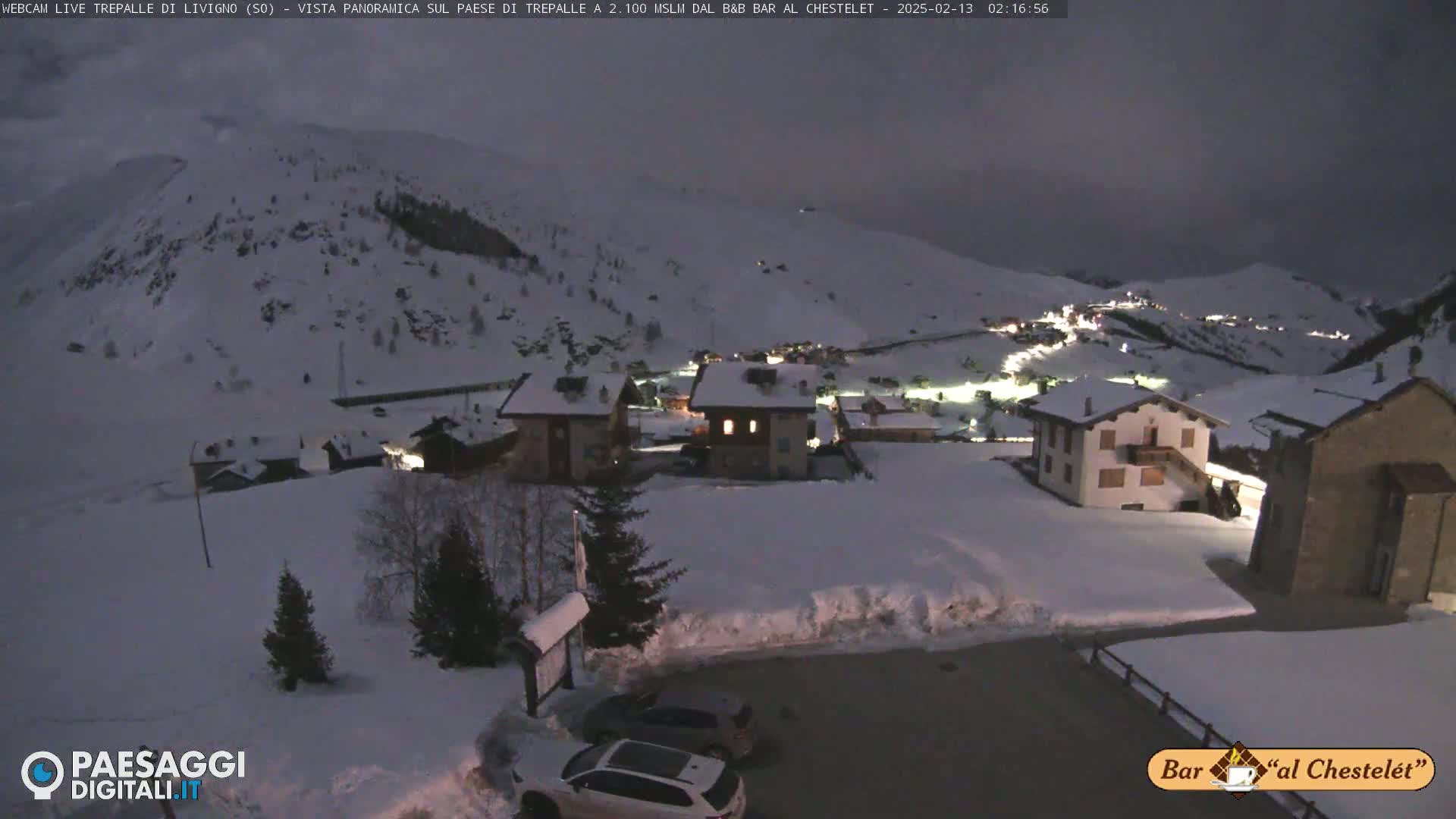 A snow-covered village at night, illuminated by scattered lights, sits nestled in a valley surrounded by snow-dusted mountains under a dark sky.