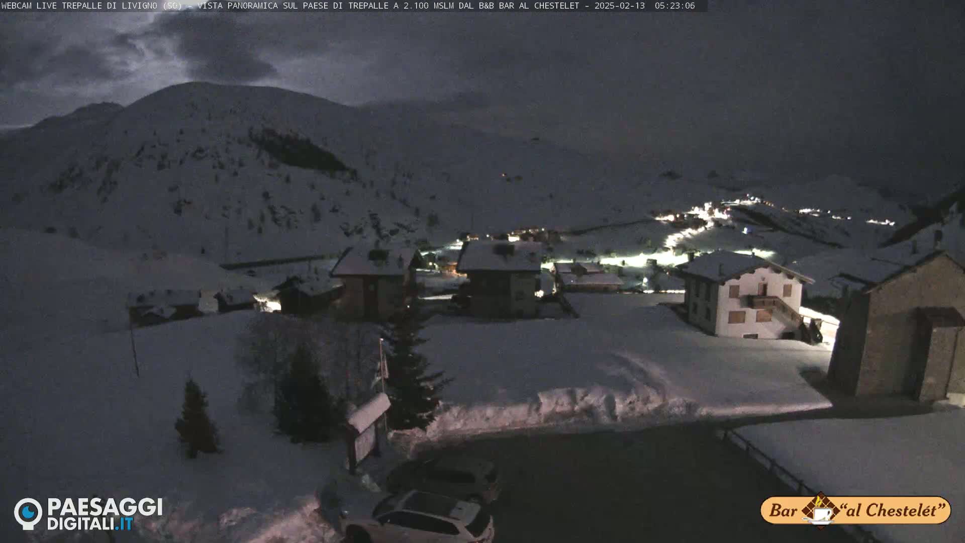 A snow-covered village at night is illuminated by scattered lights, under a dark, cloudy sky.