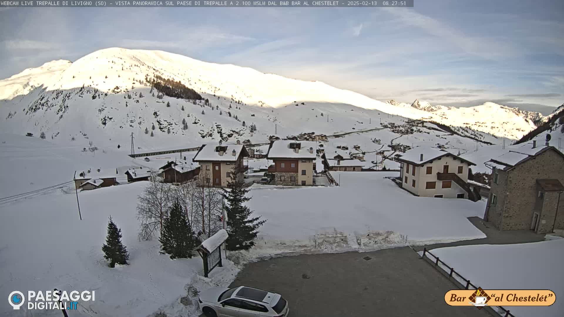 A snow-covered village nestled in a valley, with a mountain range in the background under a partly sunny sky.