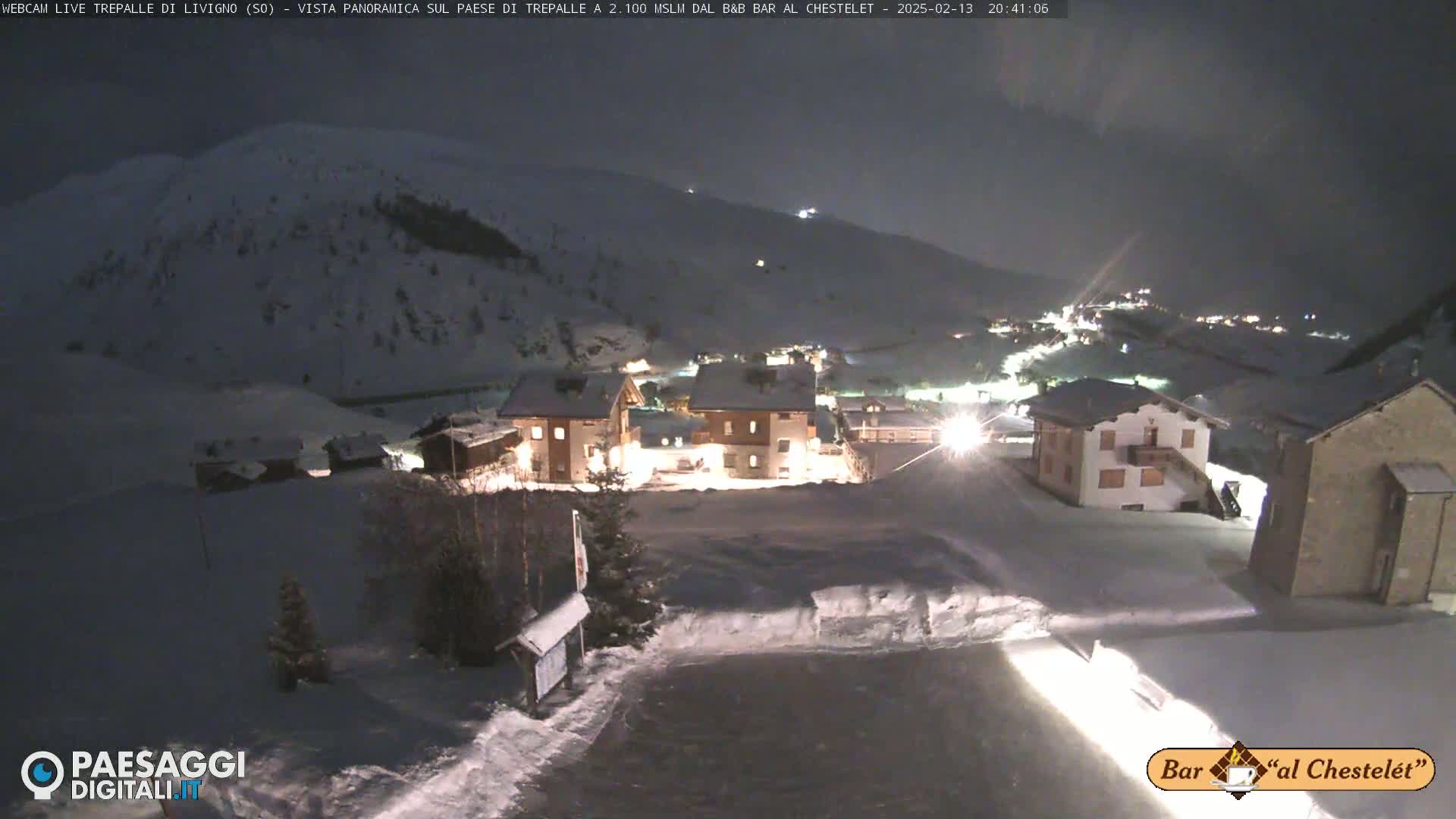 A snow-covered village is illuminated at night, nestled in a mountainous landscape under a dark sky.