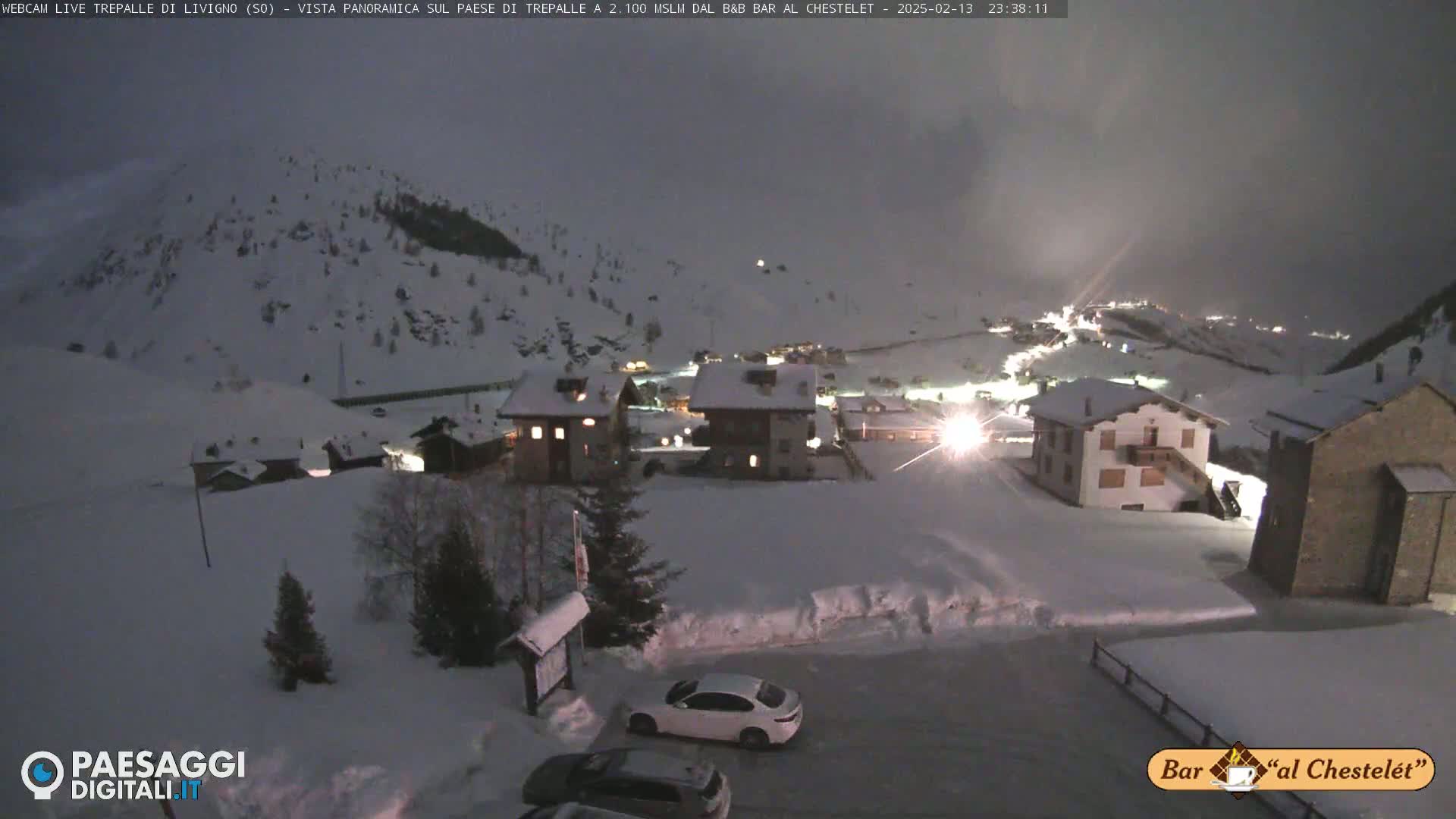 A snow-covered village at night, illuminated by lights from buildings, under a dark, cloudy sky.