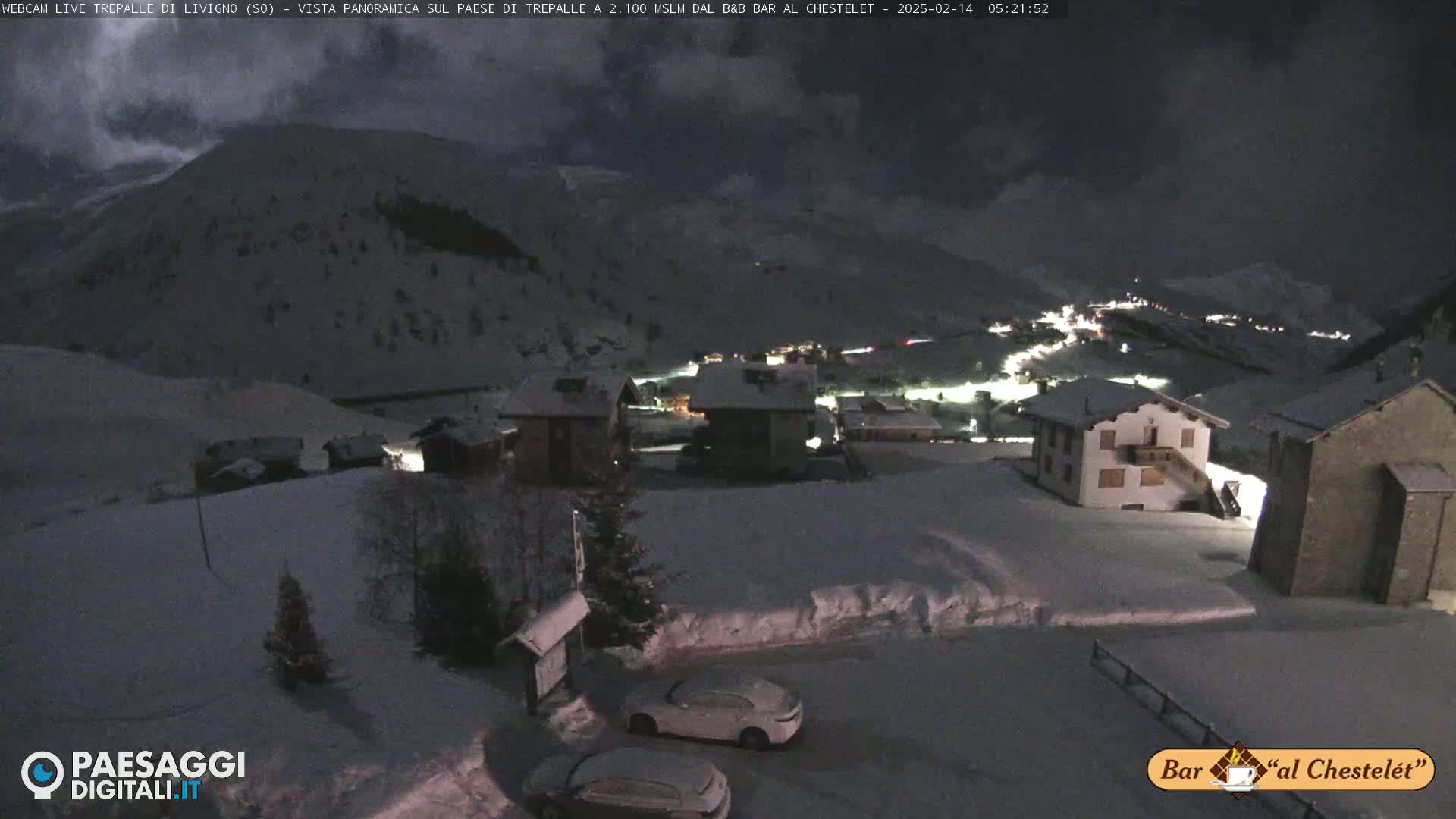 A snow-covered village at night, illuminated by scattered lights, sits nestled in a mountainous landscape under a dark, cloudy sky.