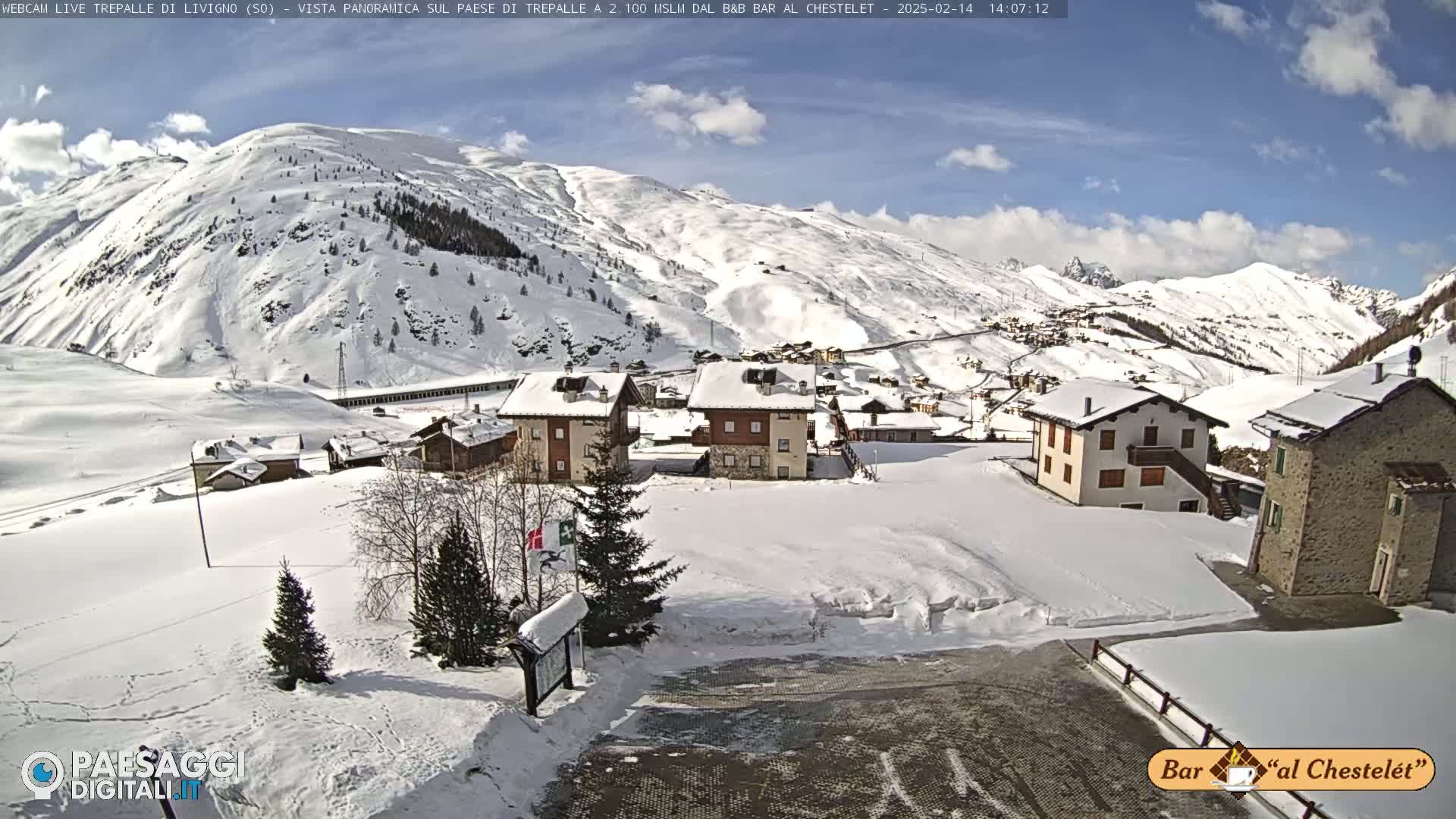 A snow-covered village nestled in a valley between snow-capped mountains under a partly cloudy blue sky.