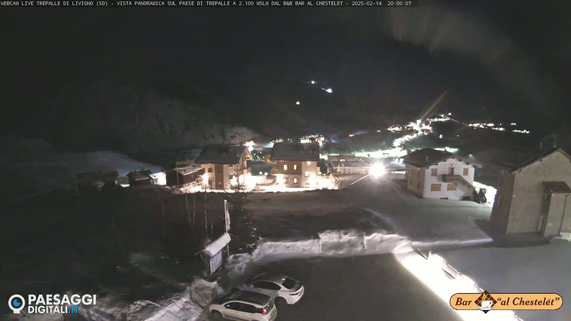 A nighttime, snowy view of a small village with illuminated buildings and a few cars parked in a snow-covered lot.