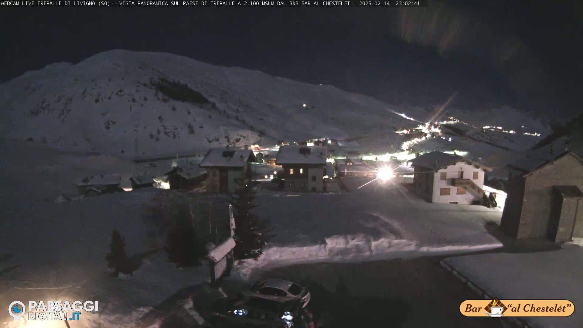 A snow-covered village at night, illuminated by lights, sits below a snow-covered mountainside under a dark sky.