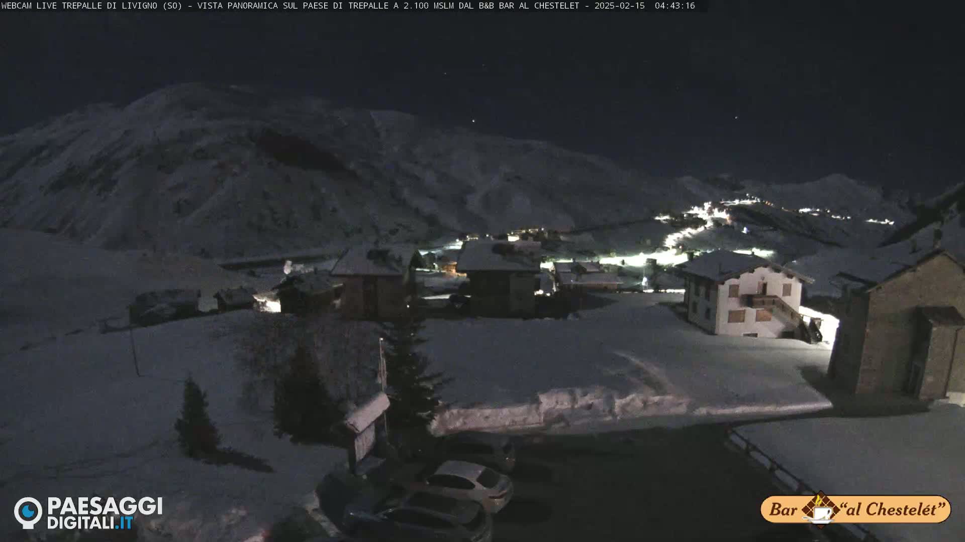 A snow-covered village is illuminated at night under a dark sky, nestled in a valley surrounded by snow-dusted mountains.