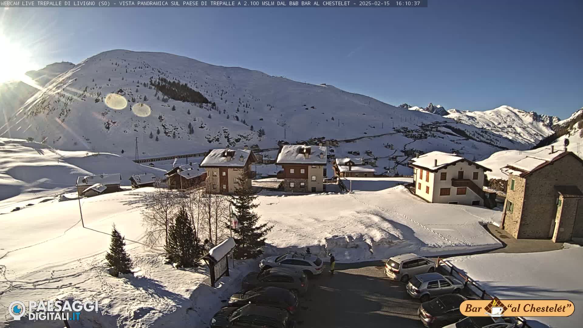 A snow-covered mountain village with several buildings, a parking lot with cars, and a sunny, clear sky.