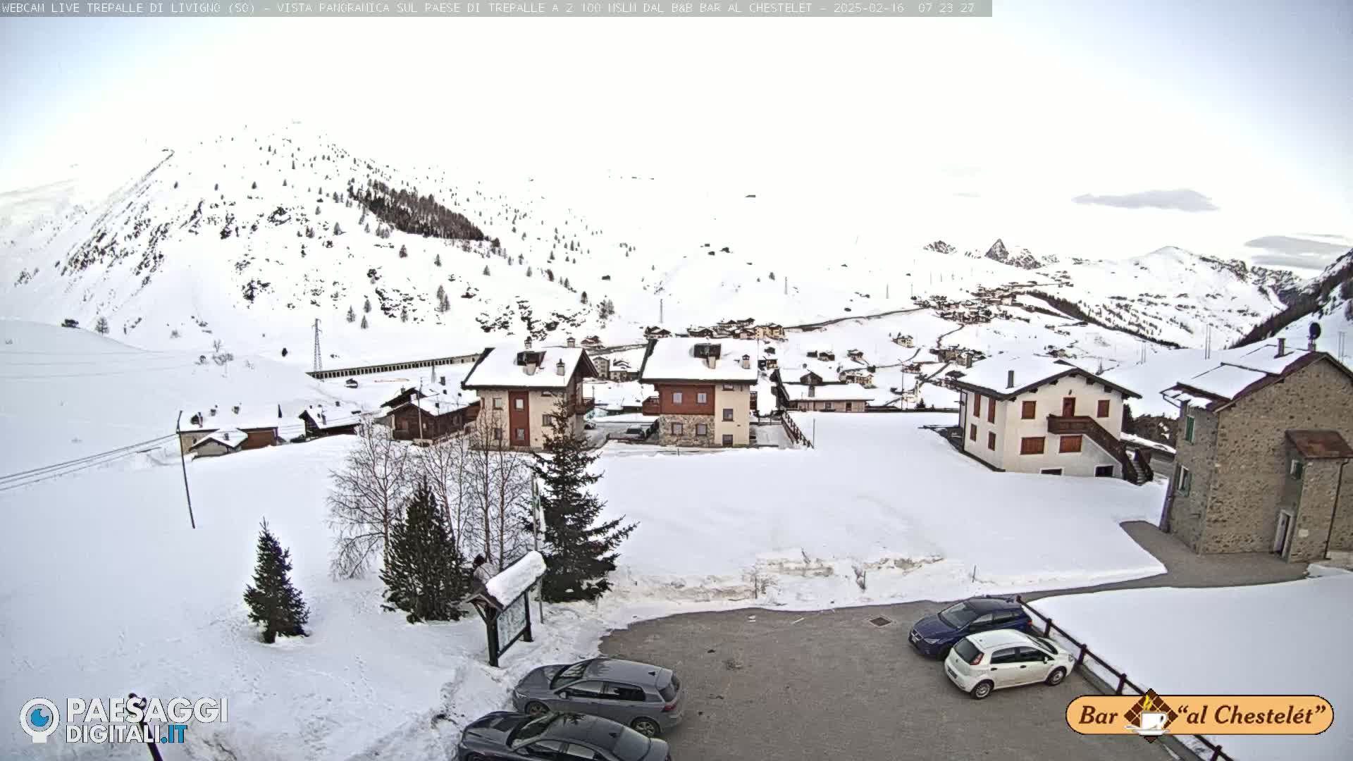 A snow-covered village nestled in a mountainous region, with several cars parked in a lot and more buildings visible in the distance under an overcast sky.