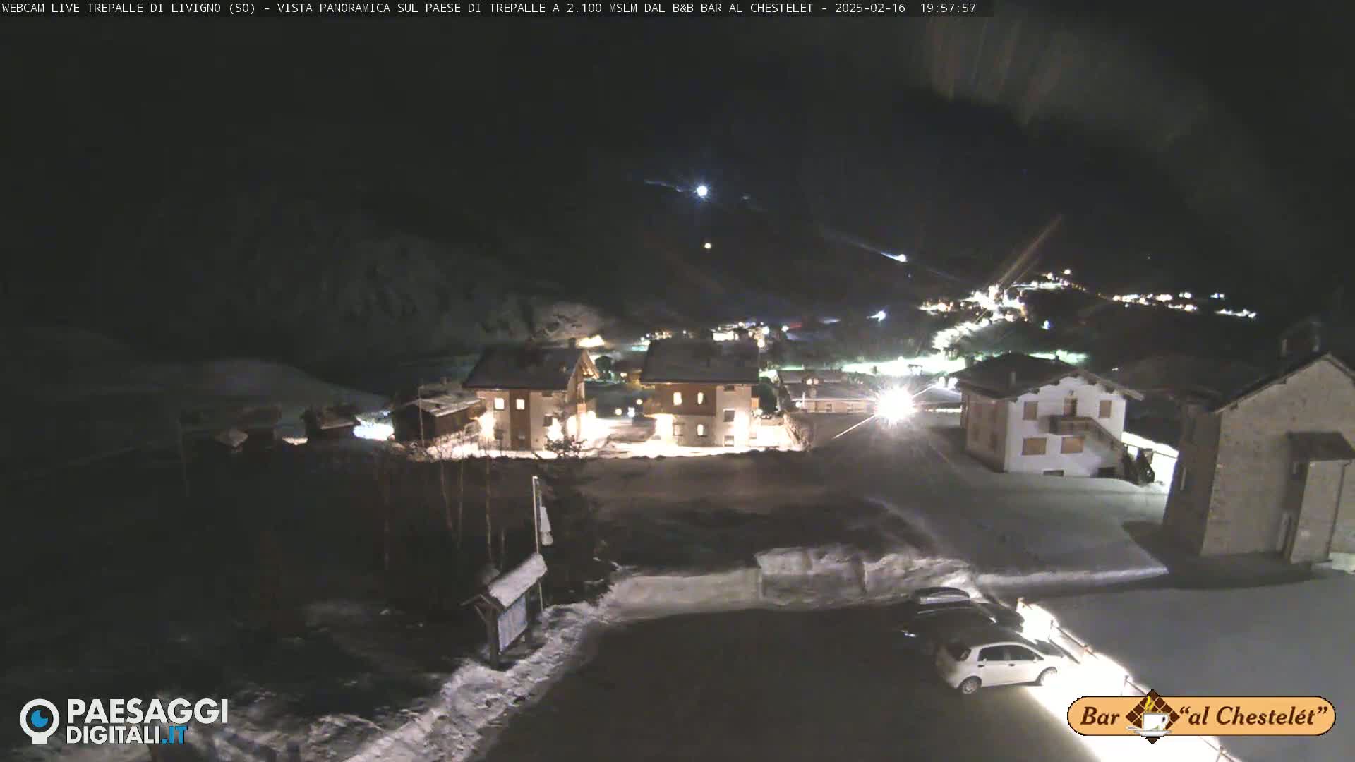 A snow-covered village at night, illuminated by streetlights and building lights, is viewed from a slightly elevated position.