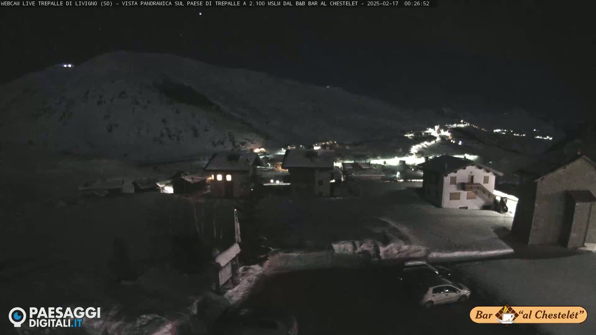 A nighttime, snow-covered village scene shows several buildings lit up under a dark sky.