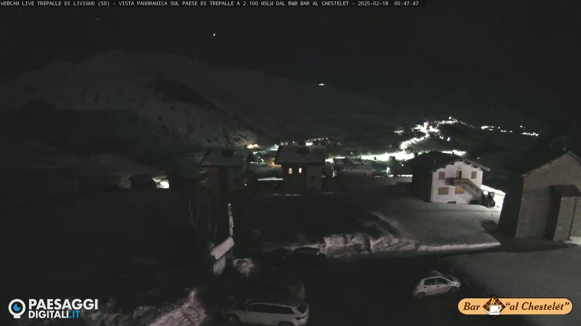 A nighttime, snow-covered village scene is visible, with a few cars parked in a lot in the foreground, and a mountainous landscape in the background under a clear, dark sky.