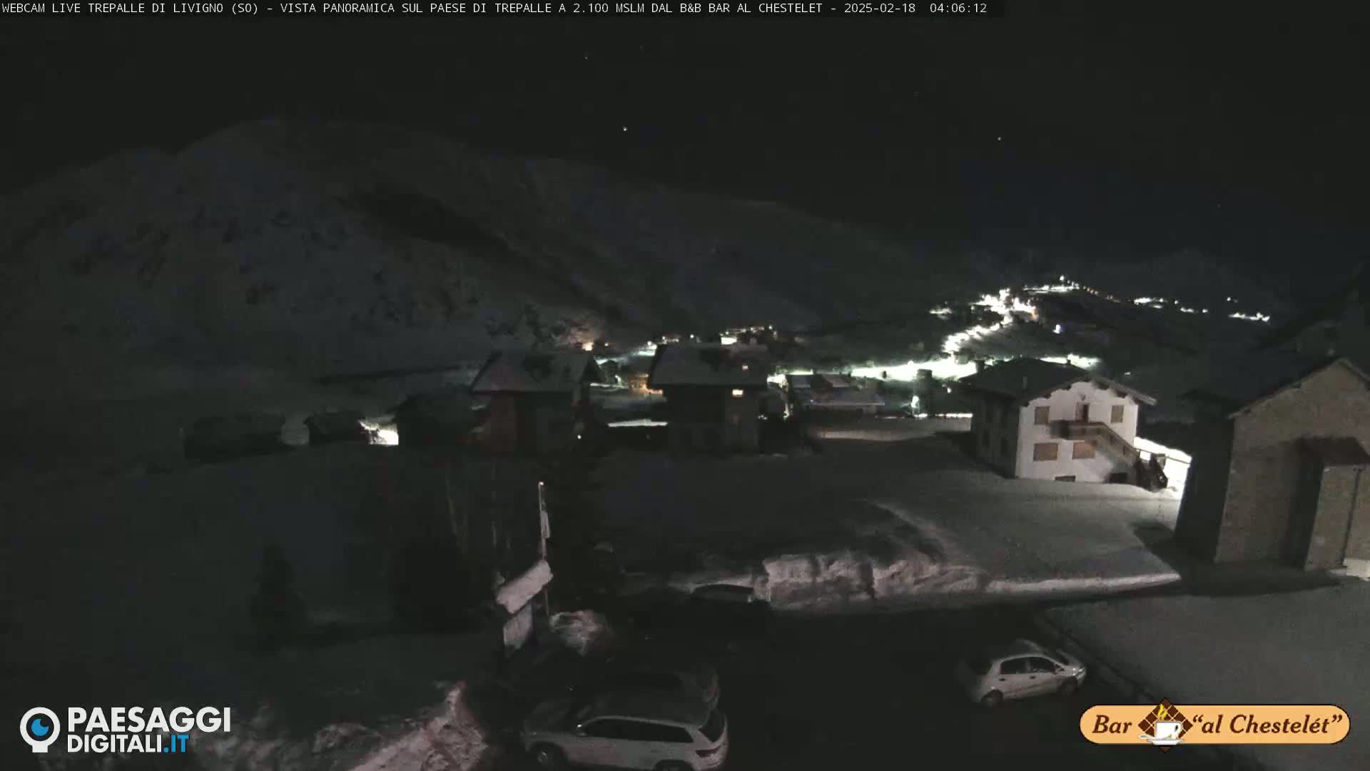 A nighttime, snow-covered village scene with a few cars parked in a lot, viewed from an elevated position under a dark sky.