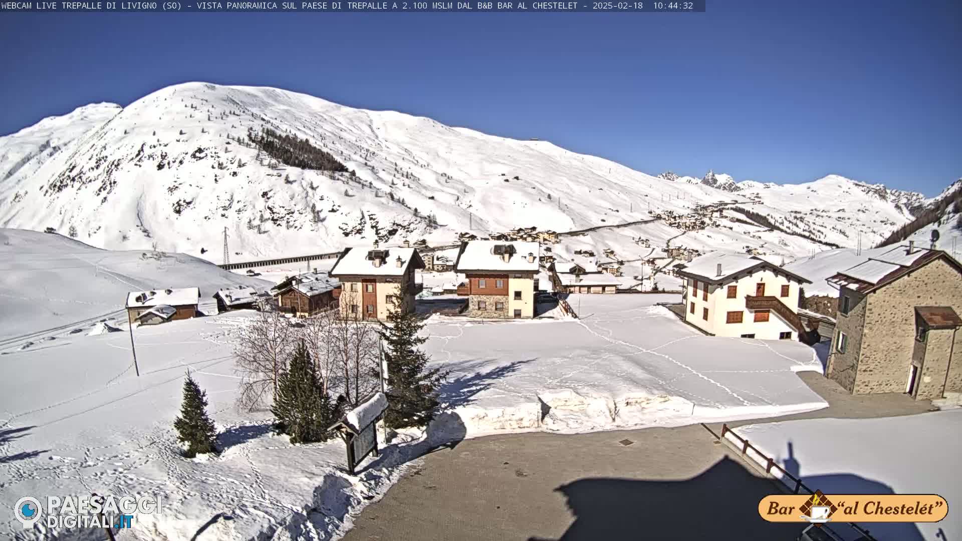 A snow-covered village nestled in a valley surrounded by snow-capped mountains under a clear, sunny sky.