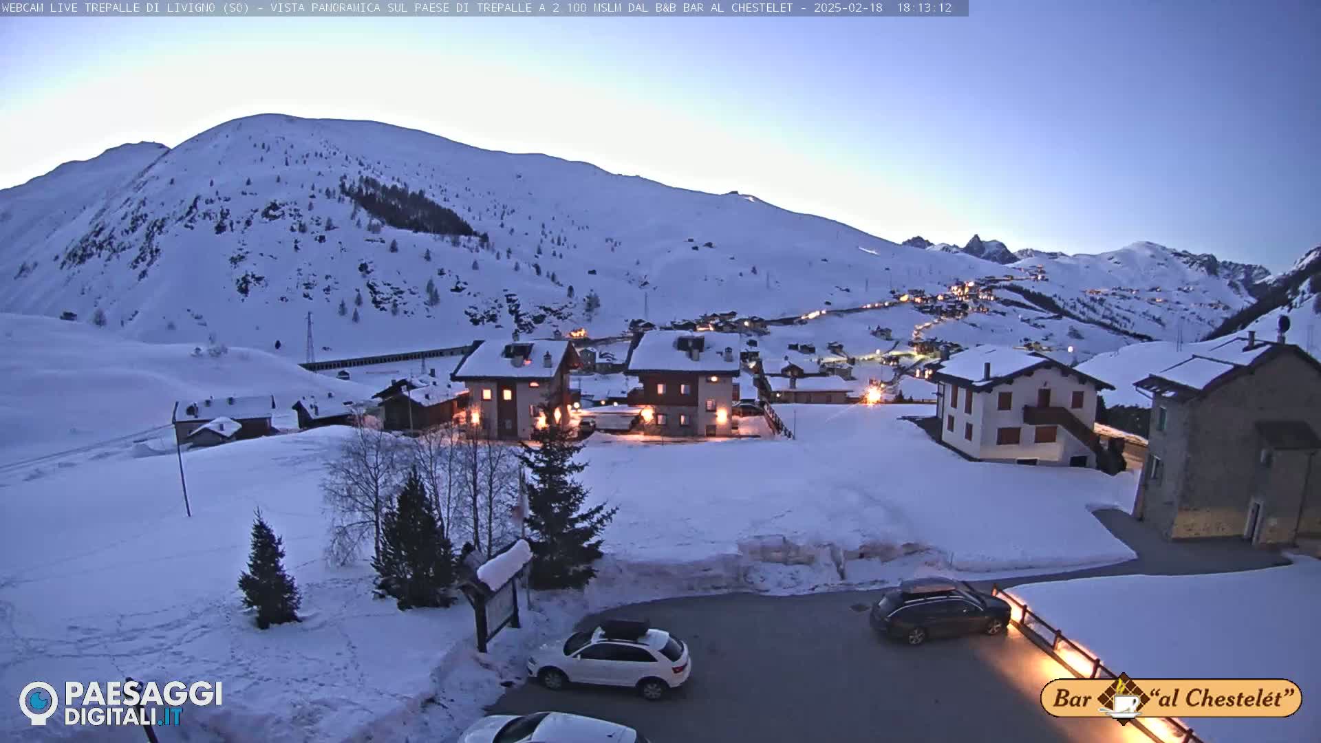 A snow-covered mountain village at twilight, with several buildings lit up and cars parked in a snow-covered lot.
