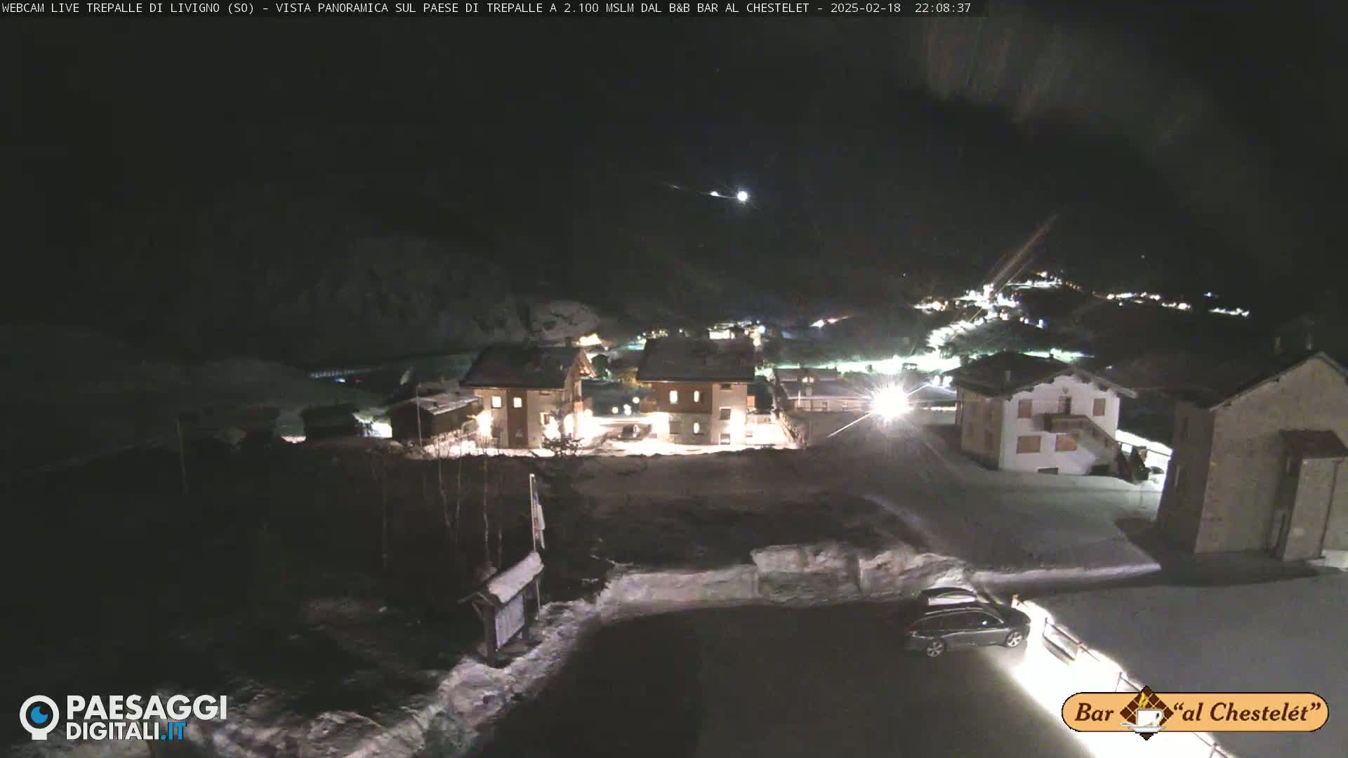 A nighttime, snow-covered village is illuminated by streetlights, with a car parked nearby.