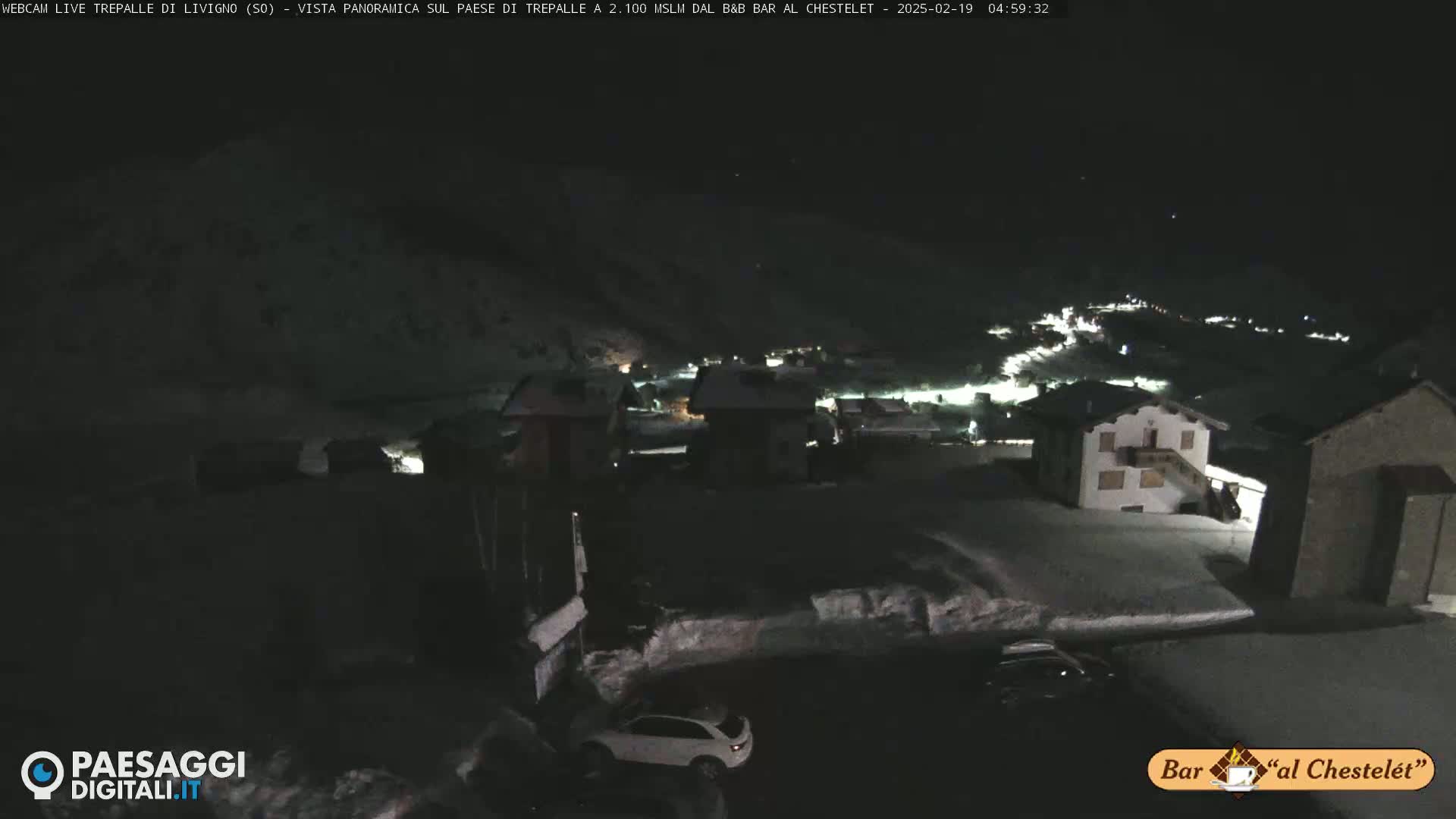 A nighttime, snow-covered village is illuminated by scattered lights under a dark sky.
