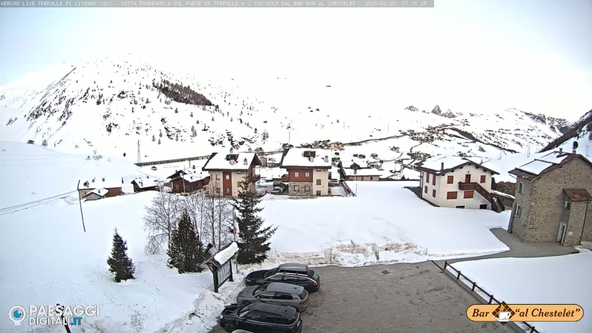 A snow-covered village nestled in a mountainous region under a cloudy sky, with several cars parked in a lot in the foreground.