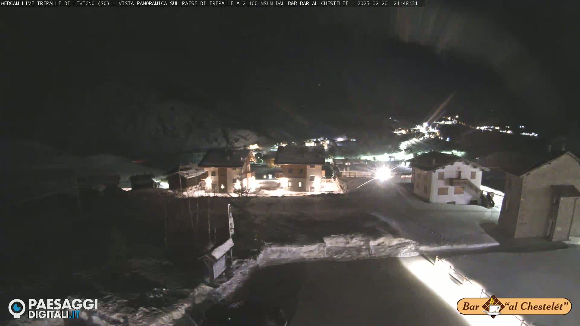 A nighttime, snow-covered village is illuminated by lights from buildings, under a dark sky.
