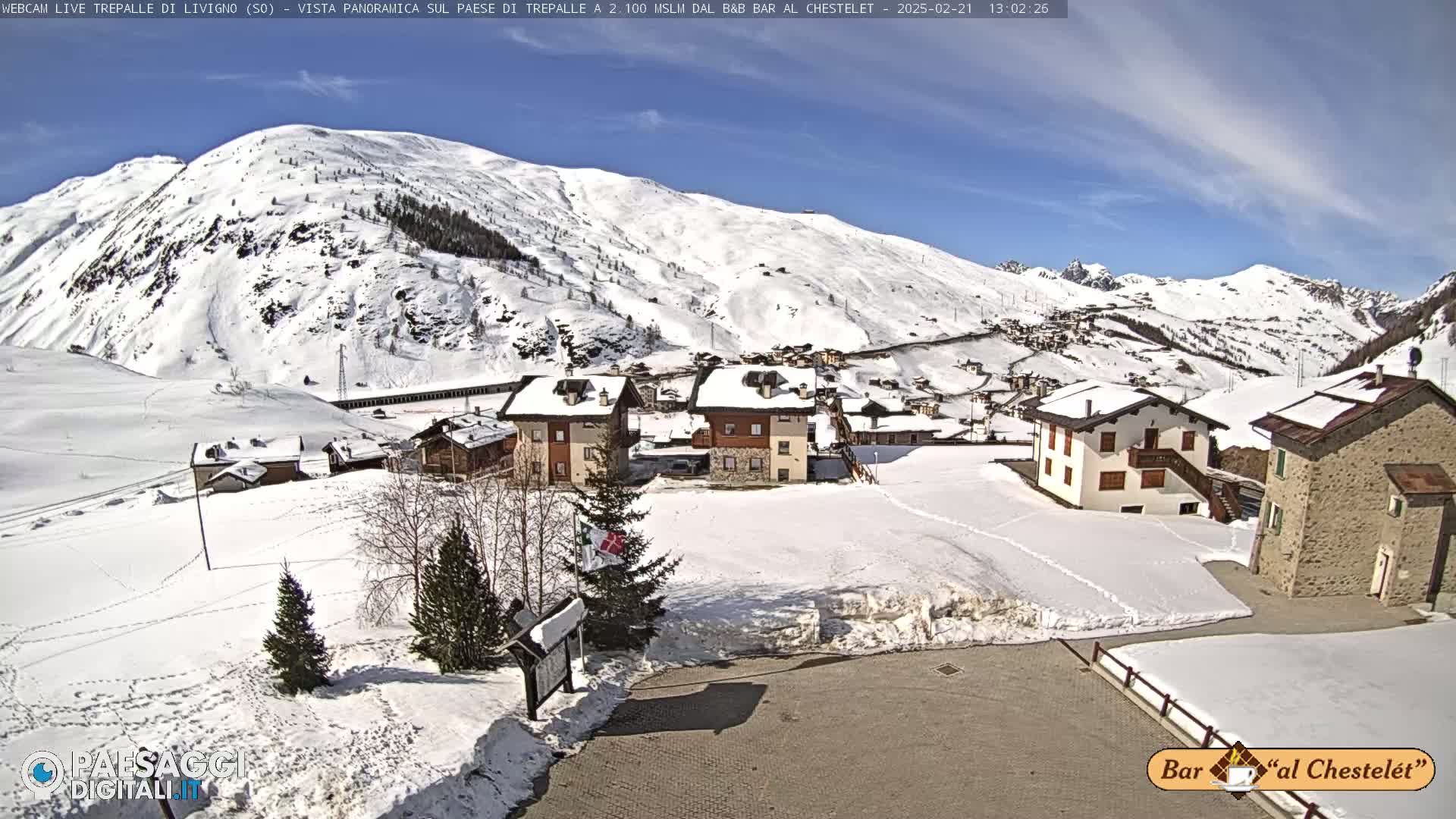 A snow-covered village nestled in a valley, with a sunny, clear sky and snow-capped mountains in the background.