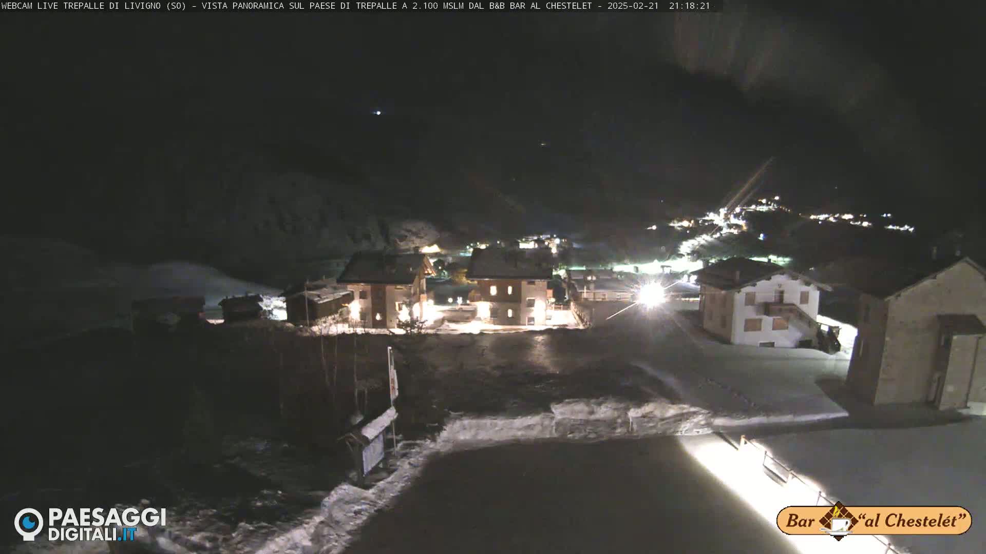 A nighttime, snow-covered village is illuminated by lights, with mountains visible in the background under a dark sky.