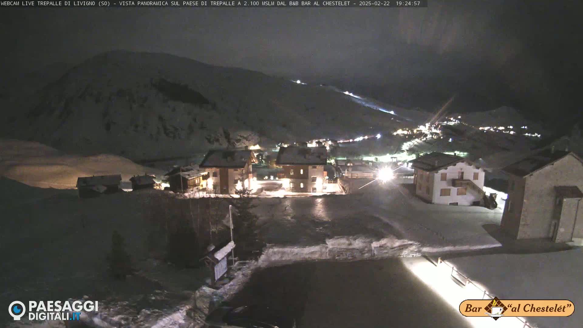 A snow-covered village at night, illuminated by lights, is nestled at the base of a snow-covered mountain.