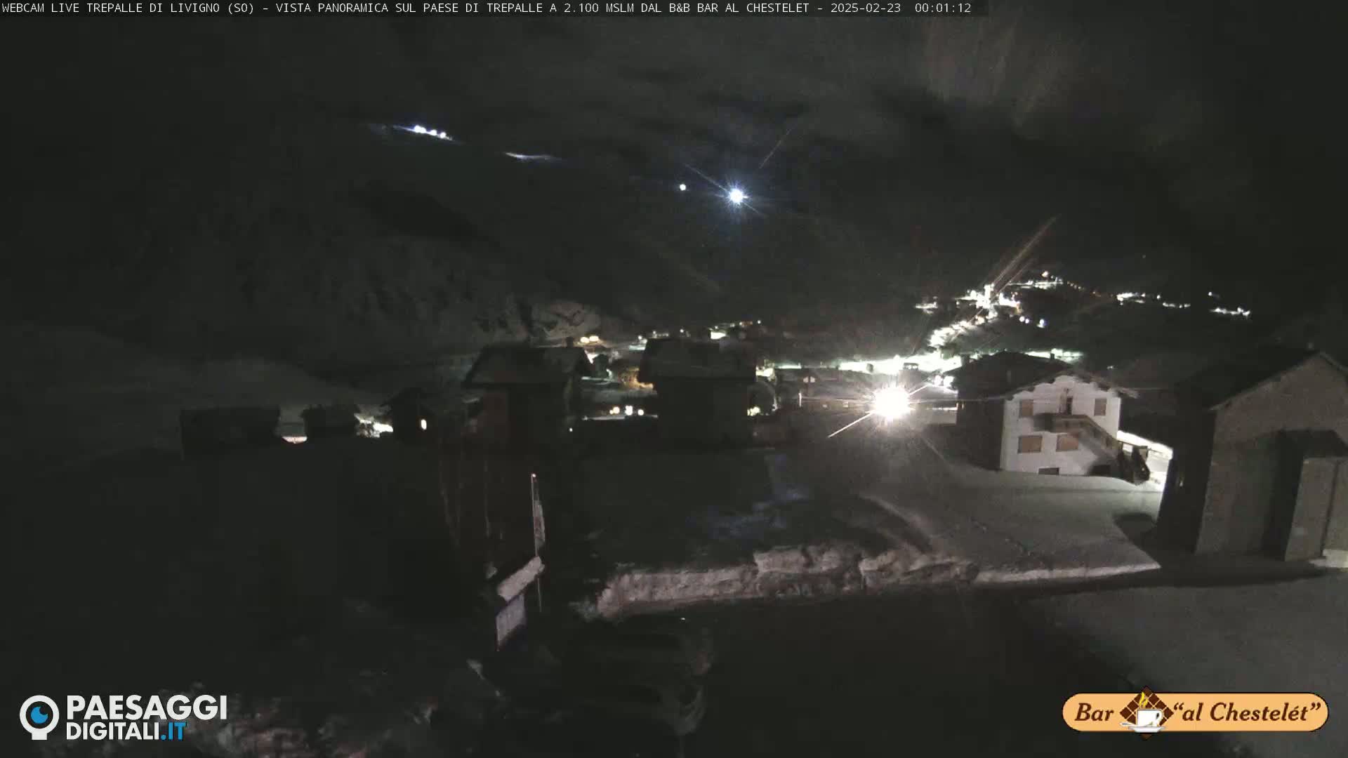 A nighttime, snow-covered village is illuminated by scattered lights under a dark sky.