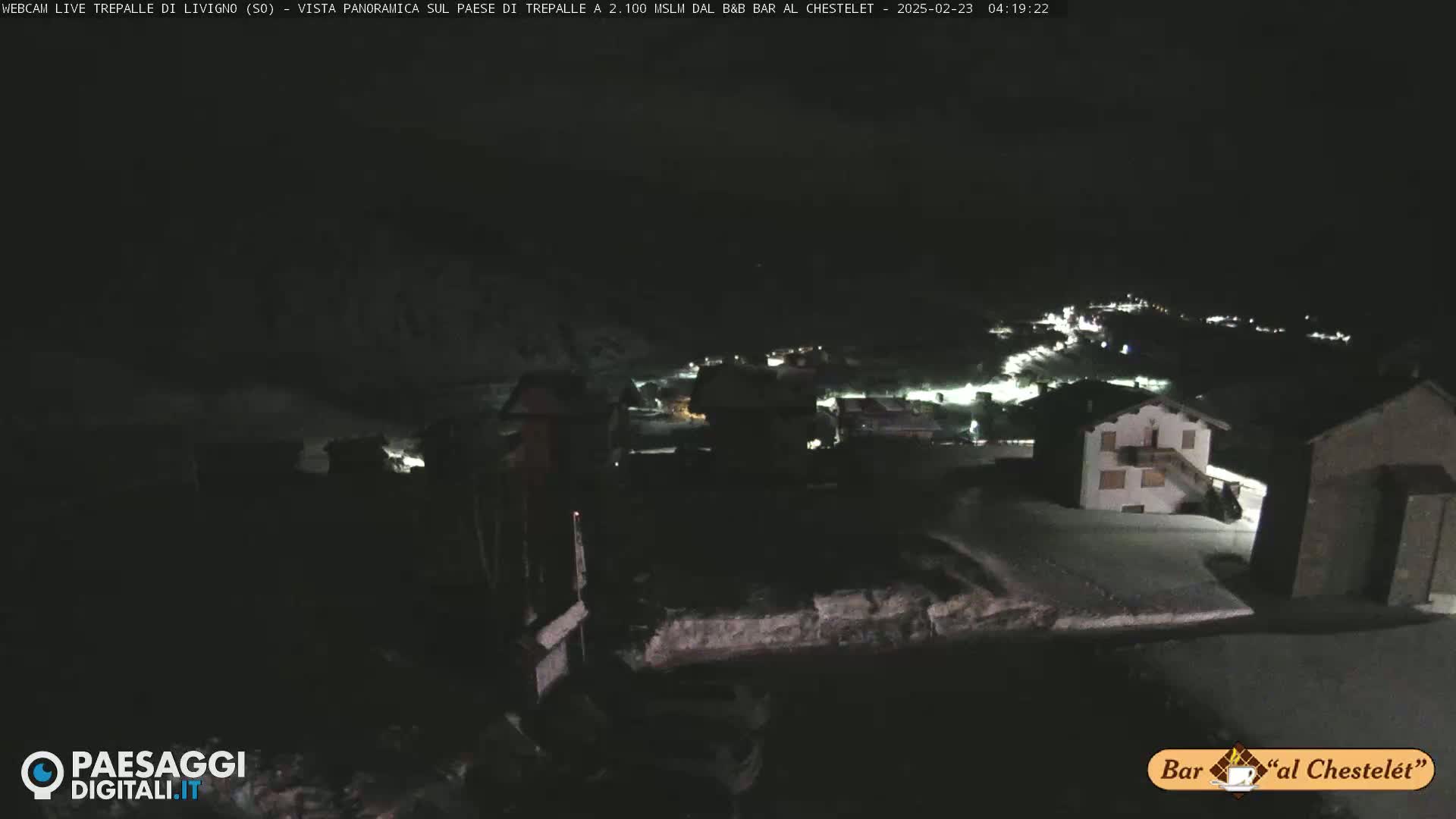 A nighttime, snow-covered village is seen from a high vantage point, under a dark sky.