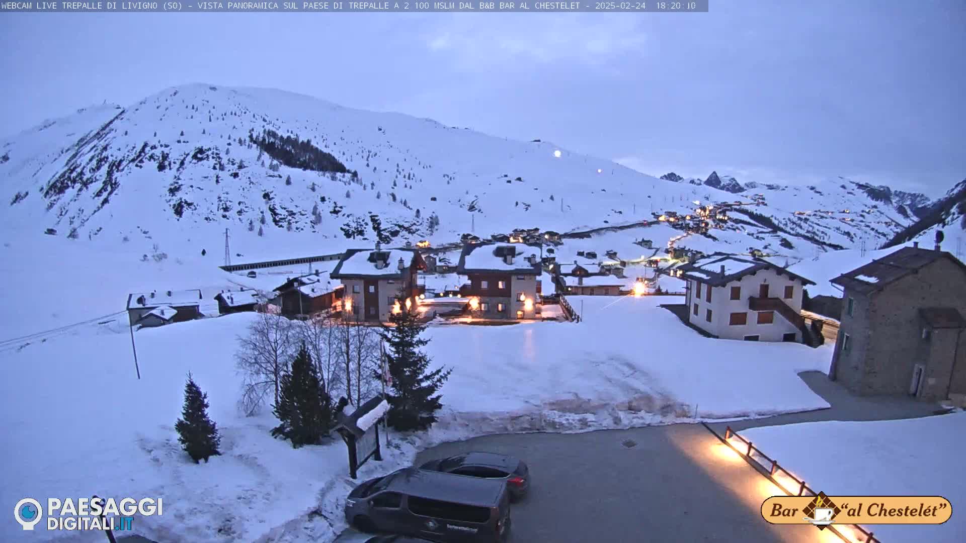 A snow-covered village in the mountains at twilight, with a few lights on in buildings and cars parked in a snowy lot.