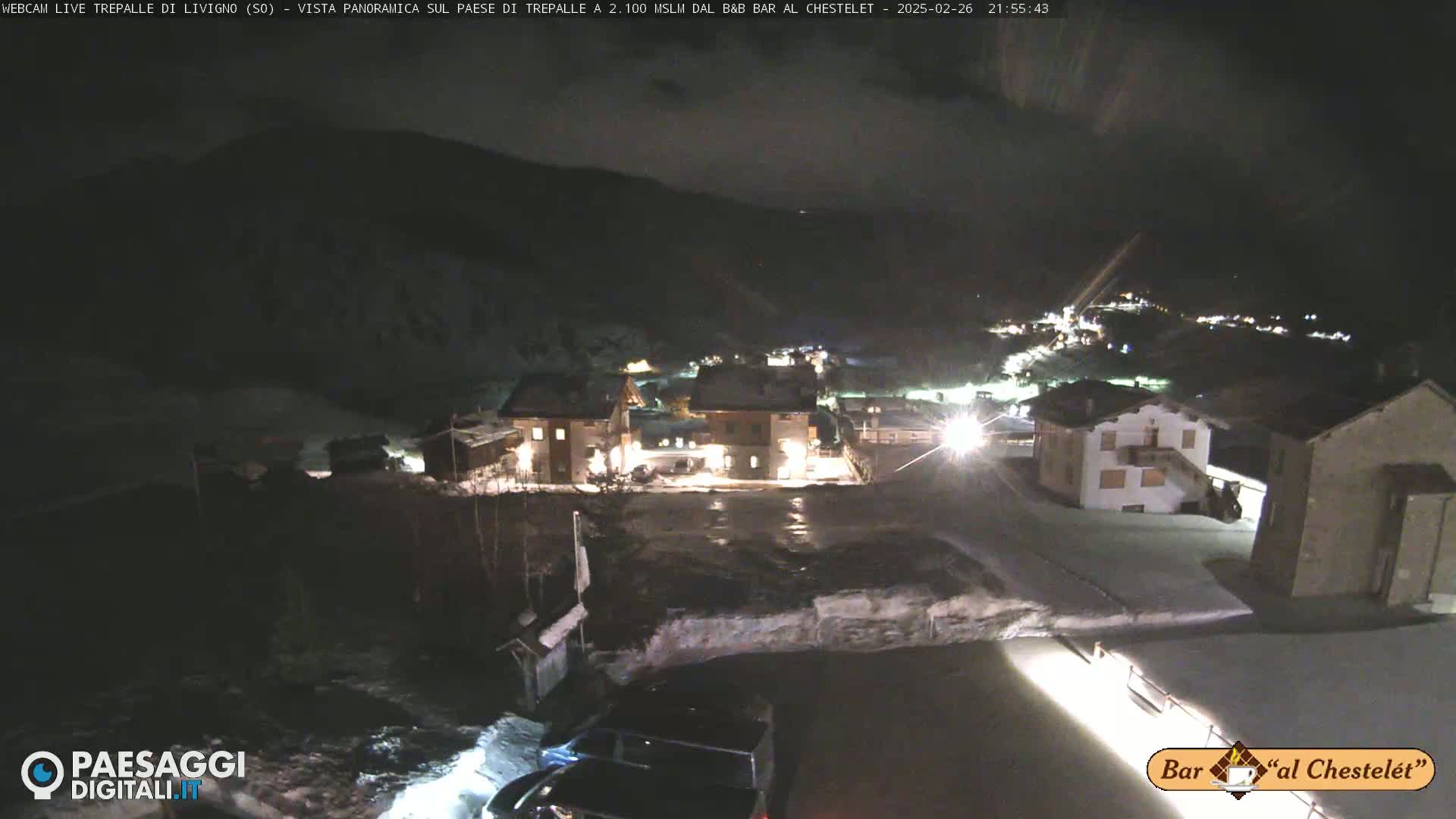 A nighttime, snowy scene shows a small village nestled in a mountain valley under a dark, cloudy sky.