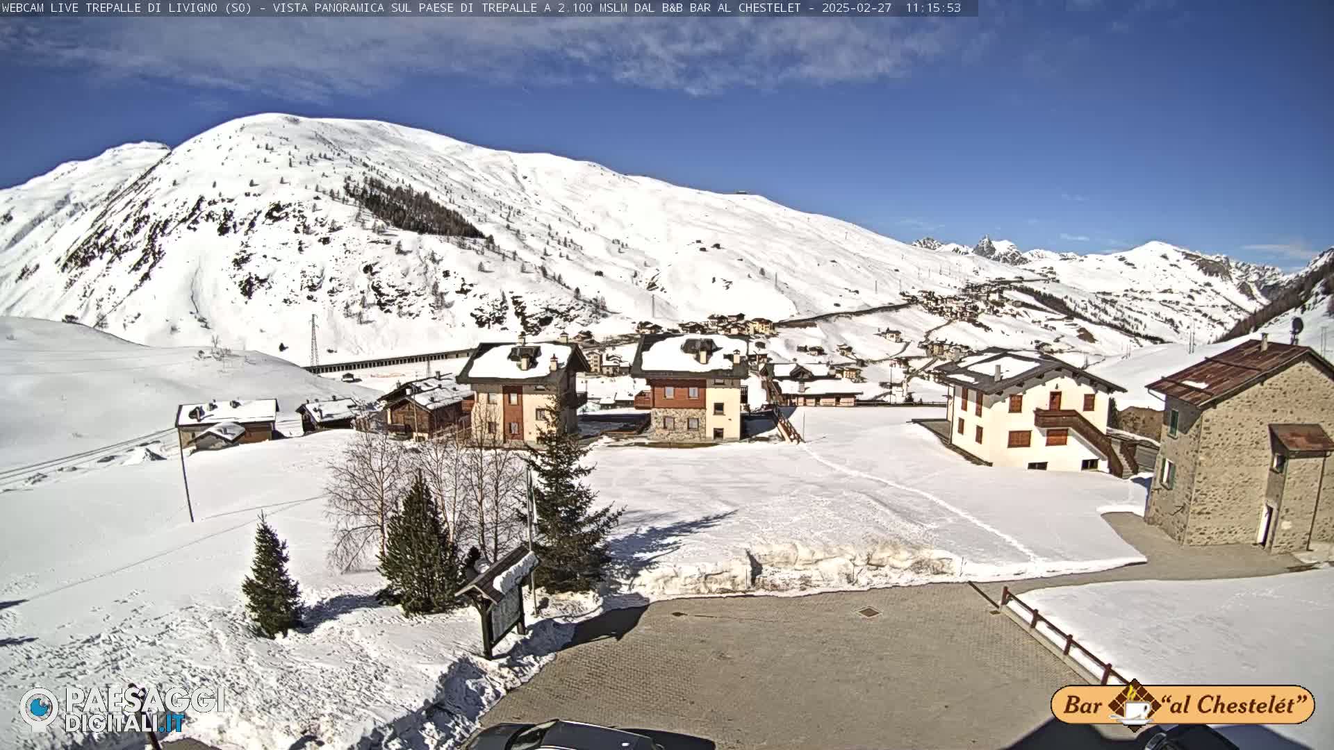 A snow-covered village nestled in a valley beneath a sunny, snow-capped mountain range.