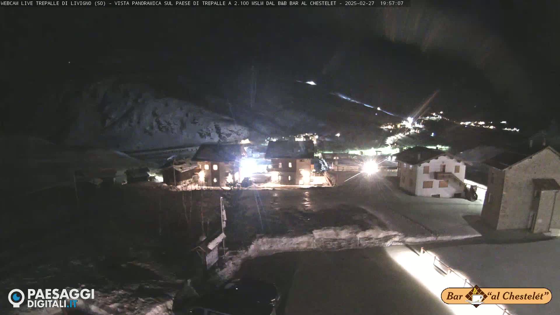 A nighttime, snow-covered village is illuminated by lights, with surrounding hills also covered in snow.