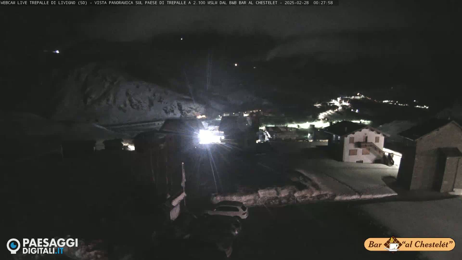 A nighttime view of a snow-covered village in the mountains under a dark, cloudy sky.