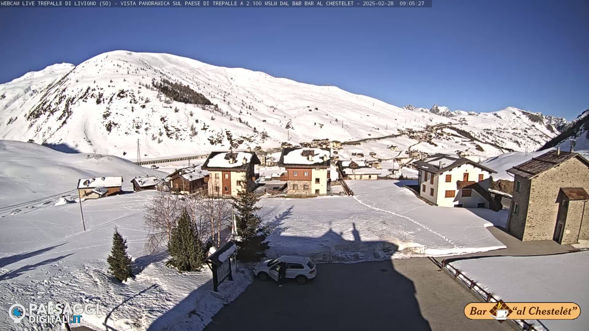 A snow-covered village nestled in a valley, surrounded by snow-capped mountains under a clear, sunny sky.