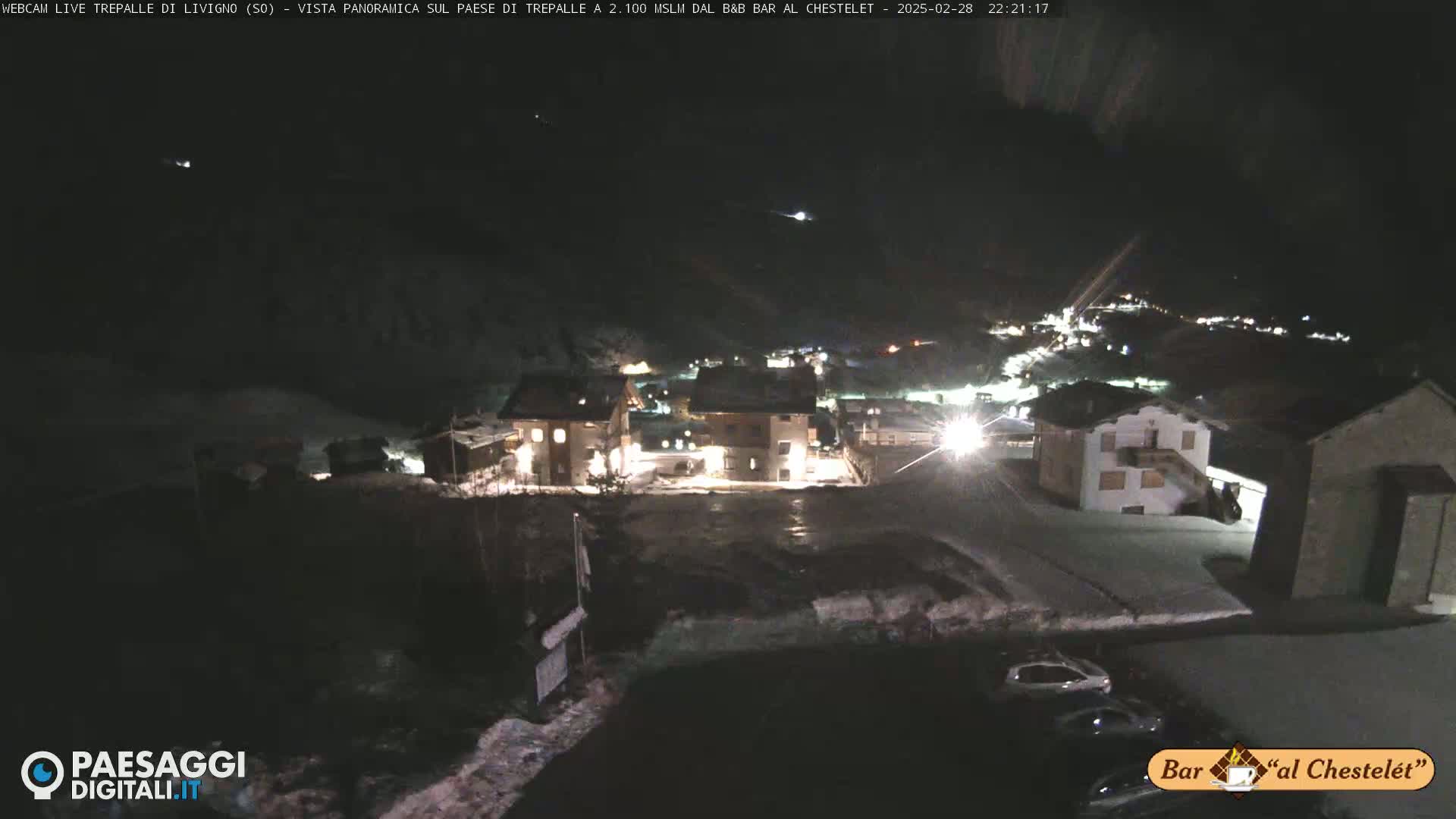 A nighttime, snow-covered village is illuminated by lights, with a few cars parked nearby.